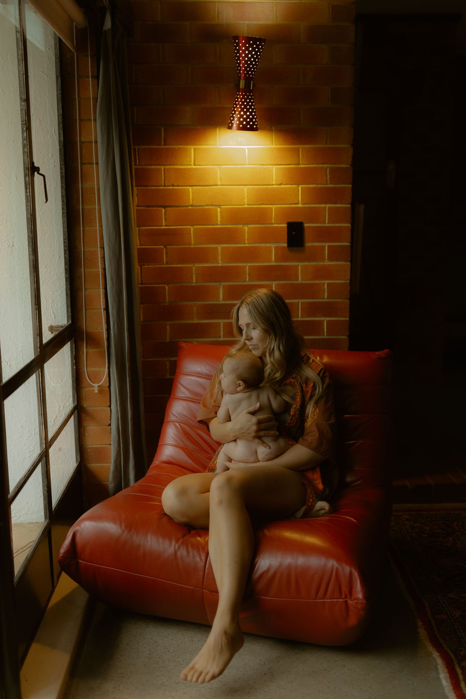 A mother sits on a red leather chair breastfeeding her newborn under warm lamp light against a brick wall.