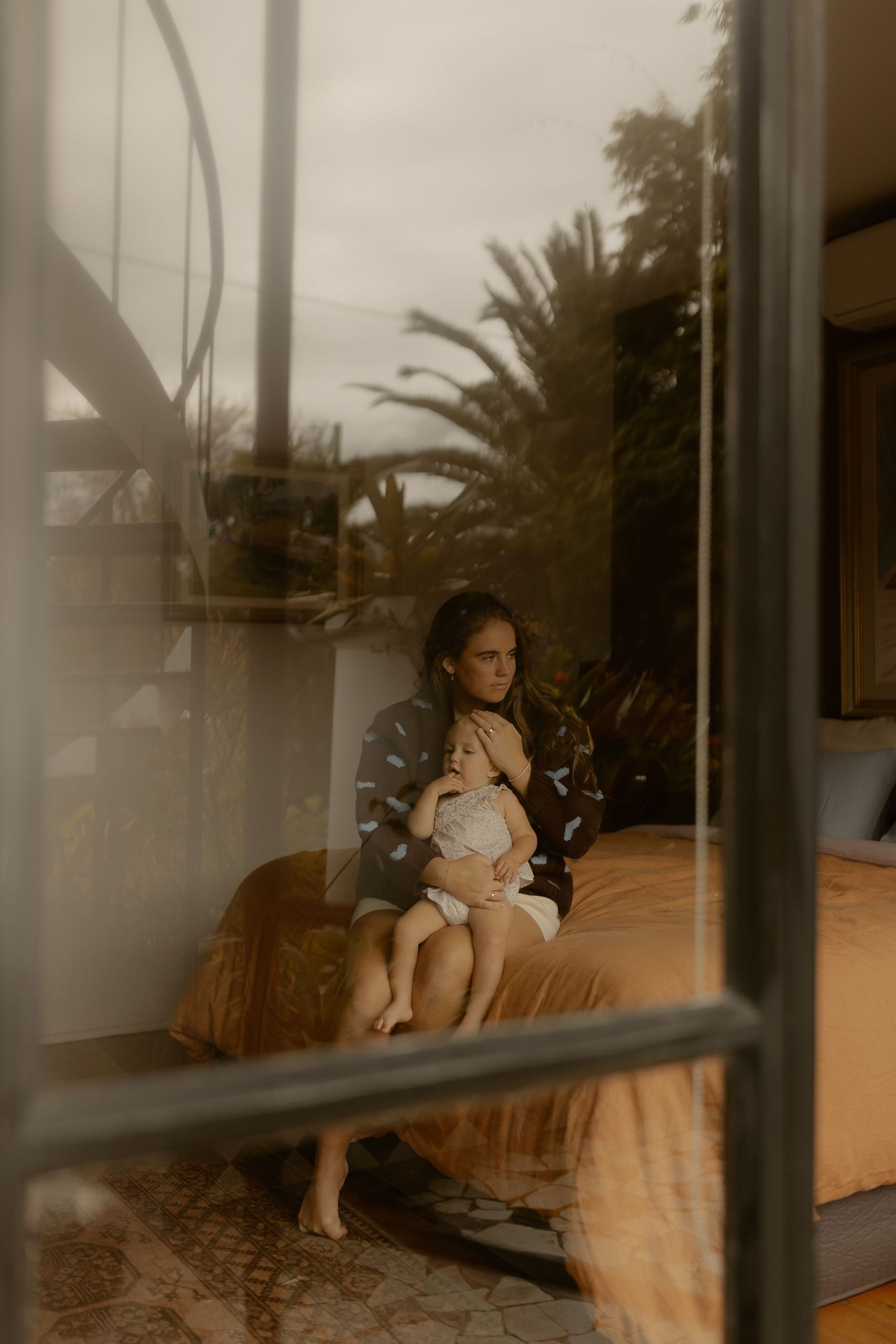 Mother cradling infant on bed photographed through window with lush palm trees in background.