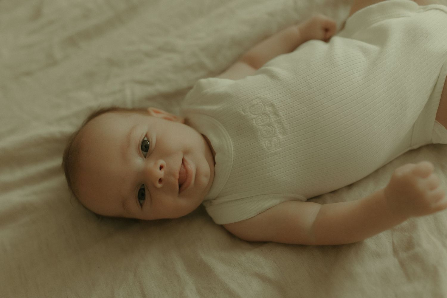 A baby in a white onesie lies on a soft blanket while looking at the camera with a gentle smile.