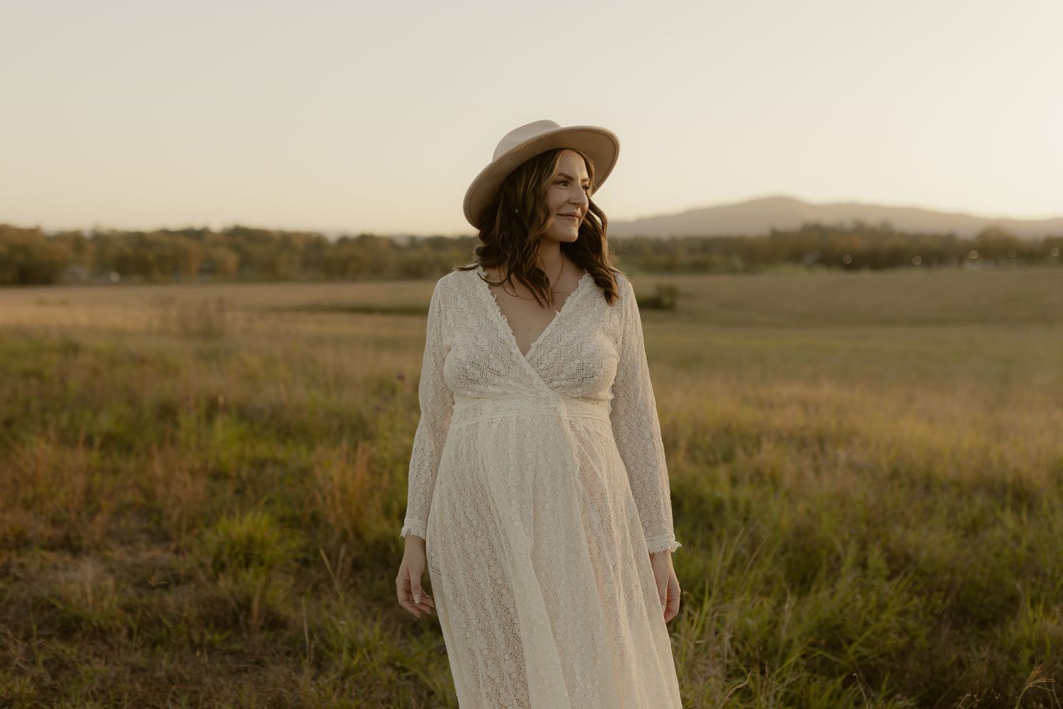 Woman in flowing white dress and hat stands in a sunlit meadow at dusk.