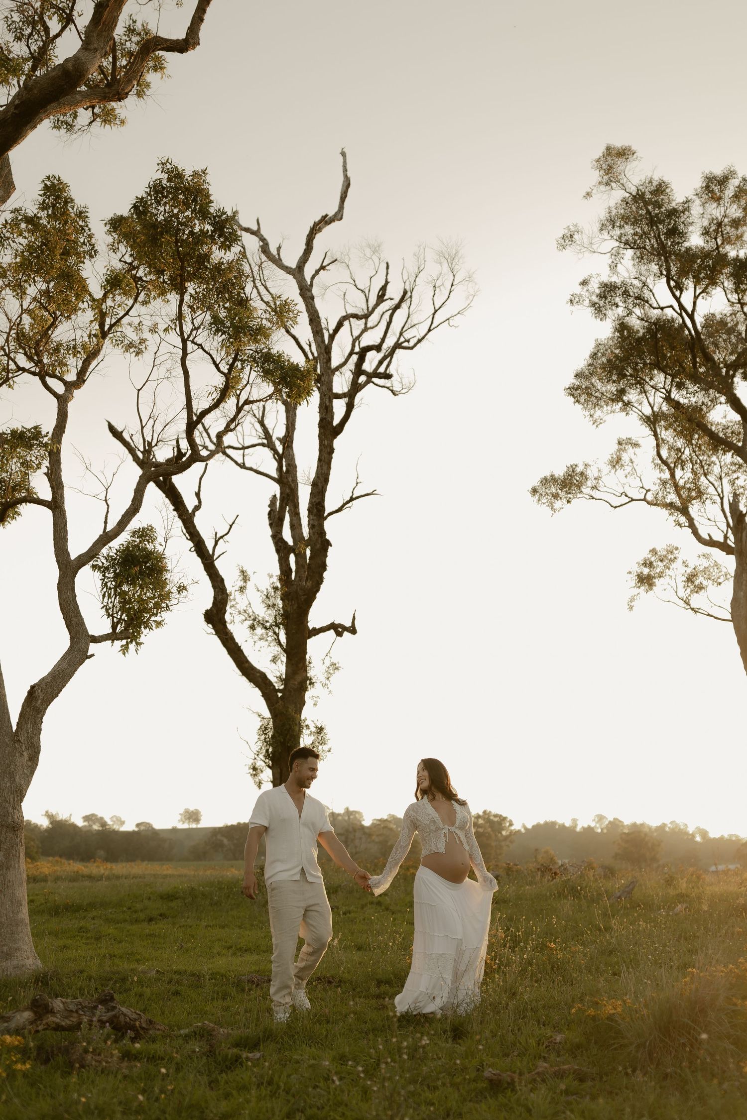 A couple walks hand in hand beneath tall eucalyptus trees during a golden sunset.