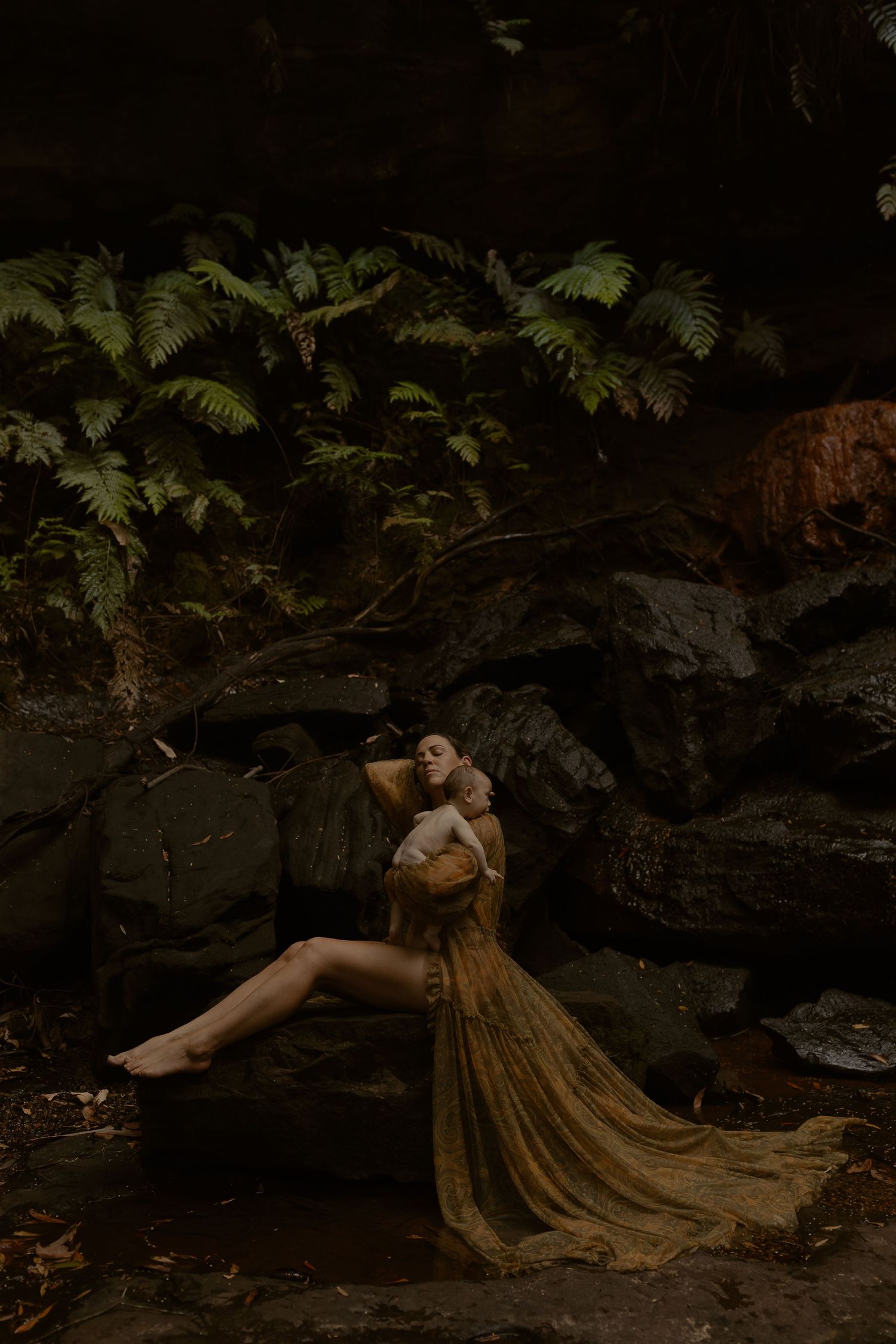 A figure in a flowing gold dress lies gracefully among dark rocks and ferns in a moody forest setting.