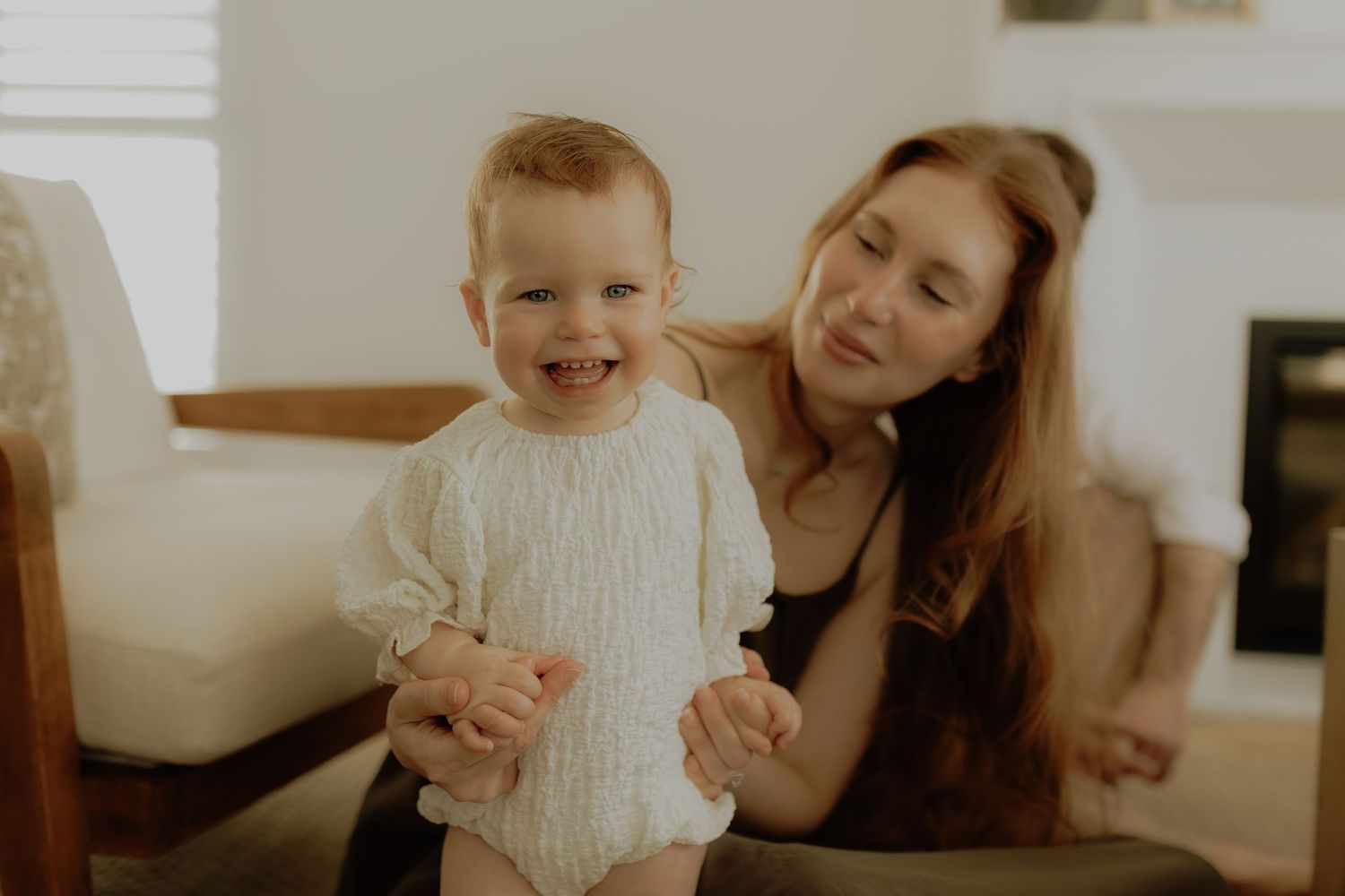 A smiling toddler in a white knit outfit stands with a young woman in a bright cozy living room.