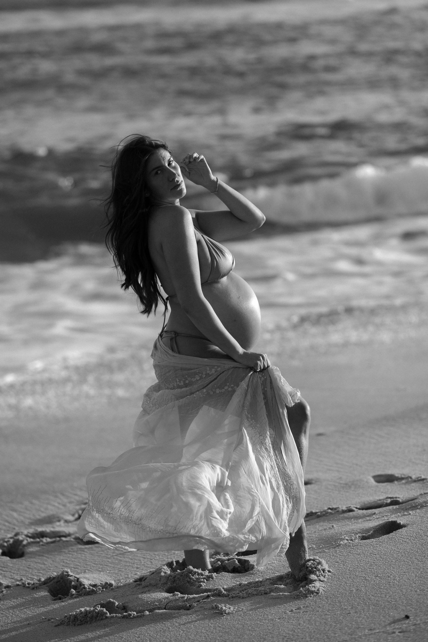 A pregnant woman in a flowing white dress poses gracefully on the beach in black and white photography.