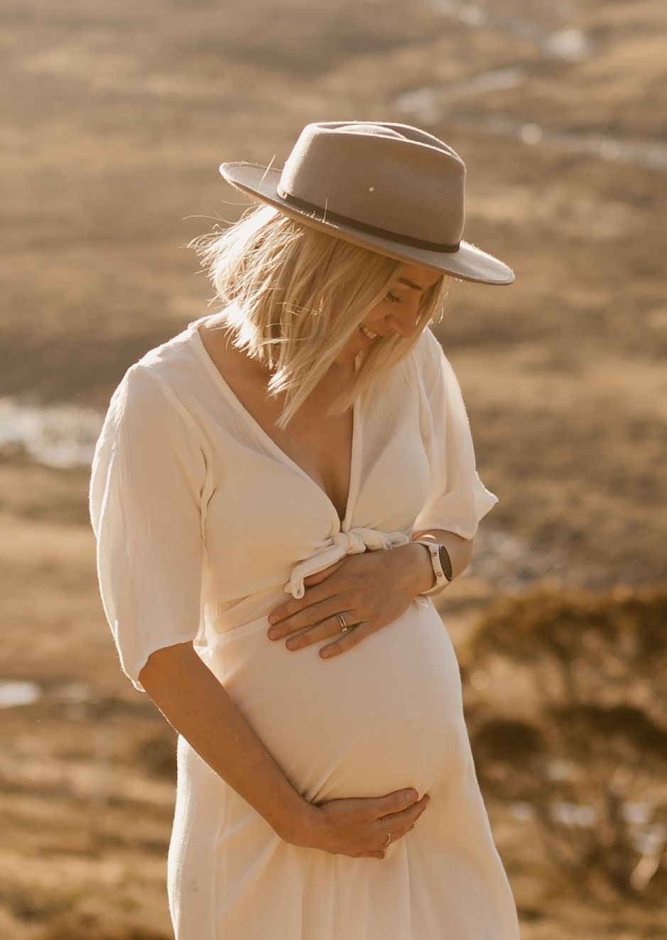 A pregnant woman in a white dress and beige hat tenderly holds her belly during a golden hour maternity photoshoot.