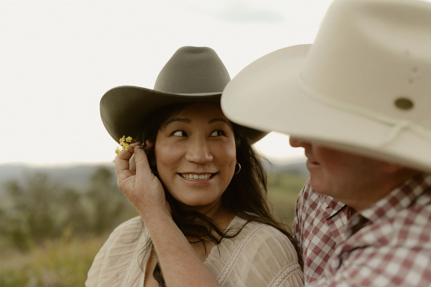 A couple in cowboy hats smiling outdoors, man placing a flower behind woman's ear in a romantic countryside setting.