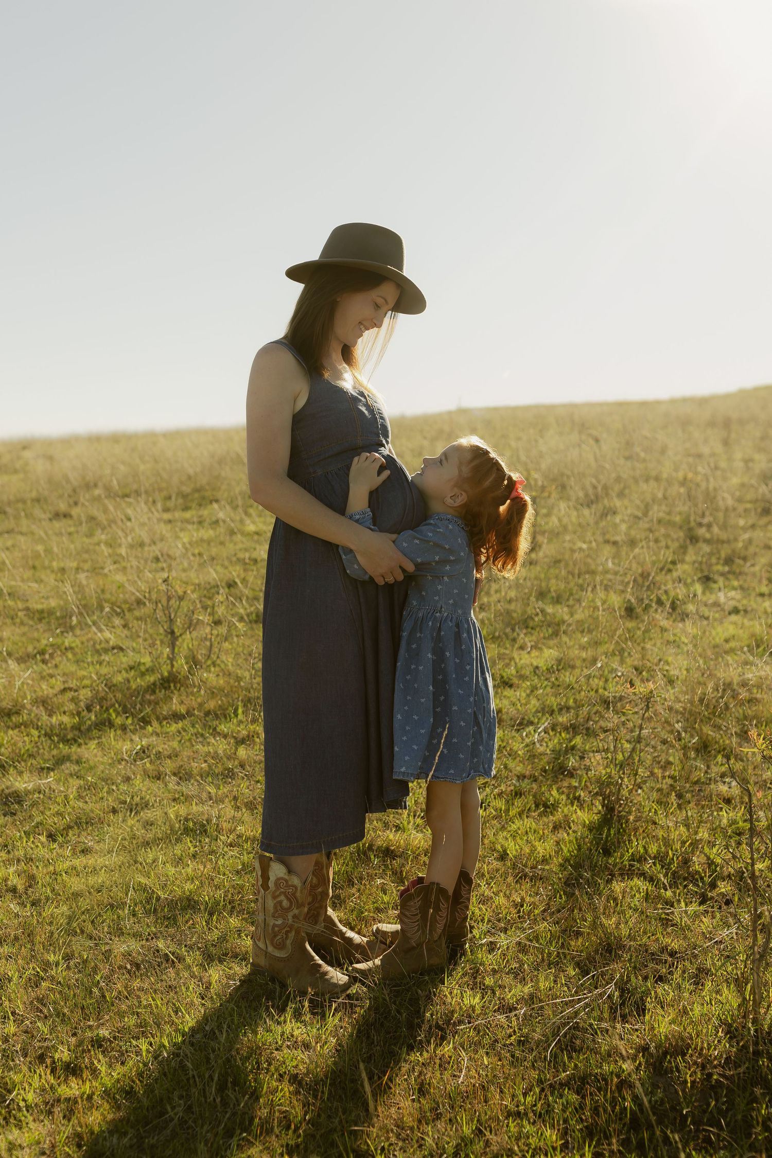 Pregnant woman in blue dress and hat with young girl in sunny green field, sharing a tender mother-daughter moment.