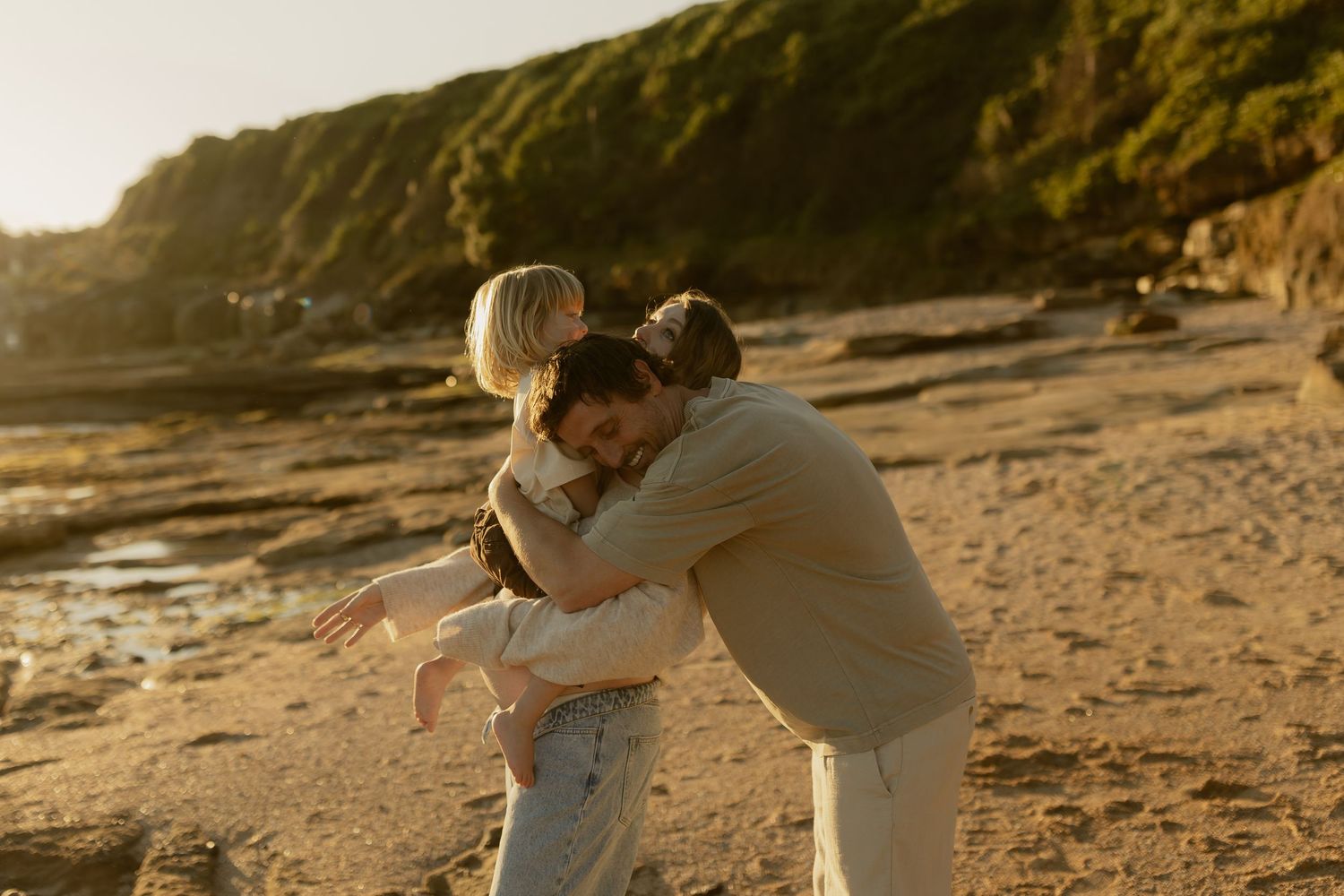Family sharing a warm embrace on a sandy beach at golden hour with green cliffs in the background.