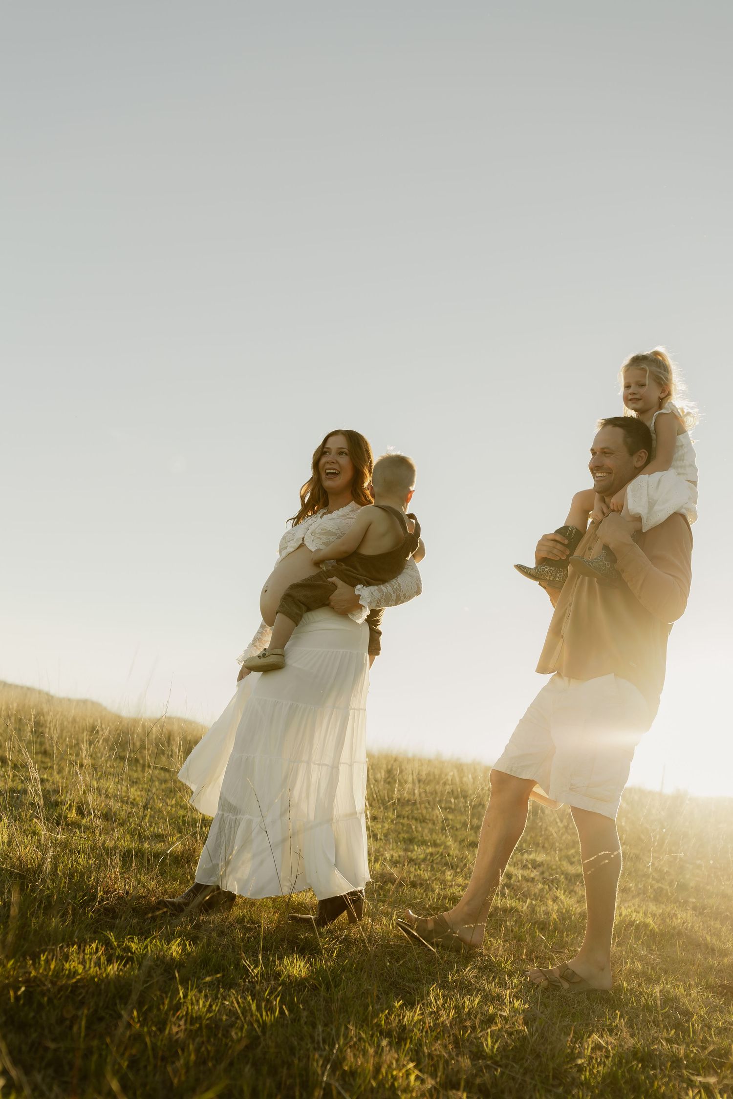 Family of four enjoying golden hour sunset on a grassy hill, mother holding baby, father with toddler on shoulders.