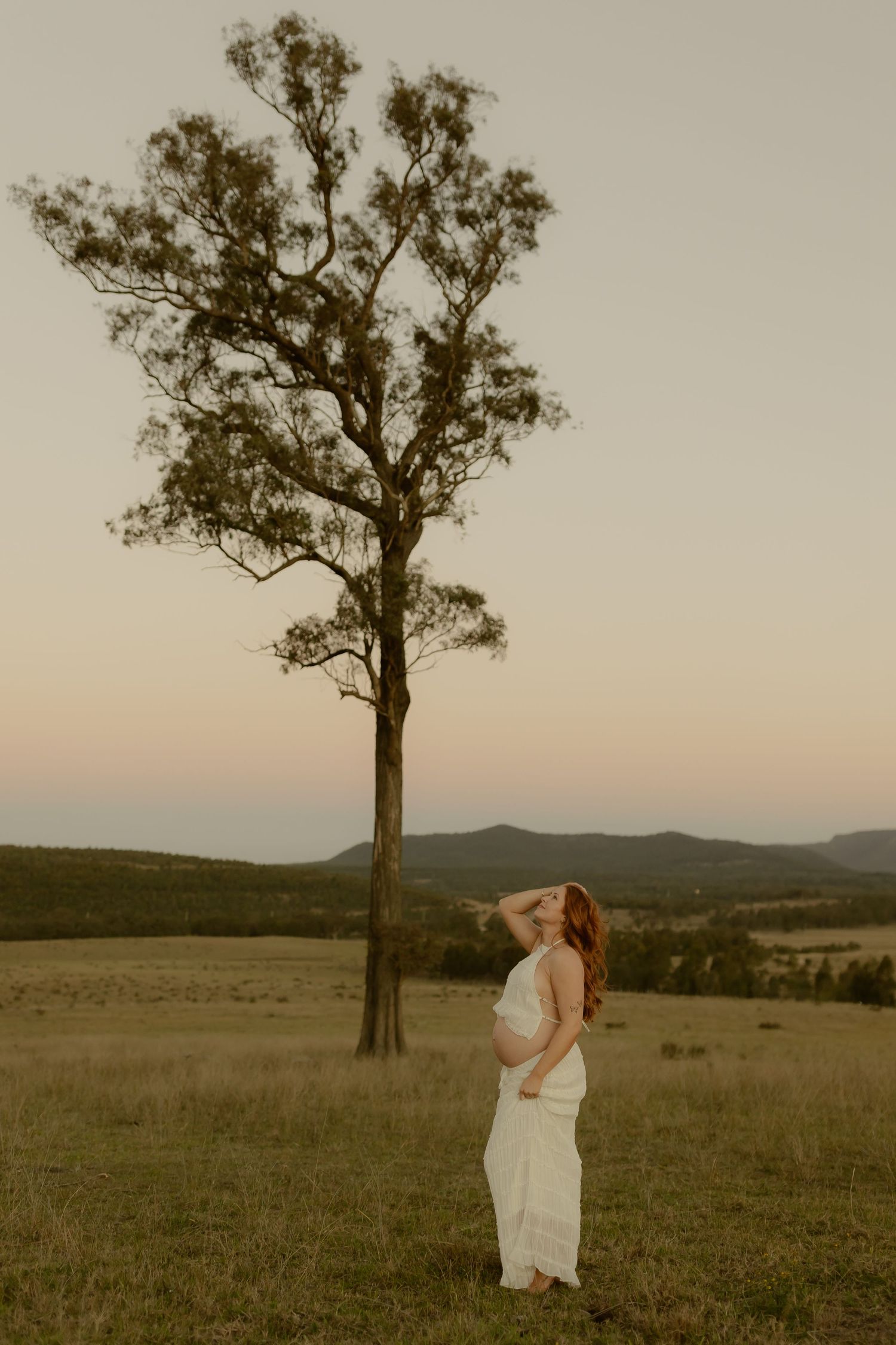 Pregnant woman in white dress standing by tall tree in golden hour field with mountain backdrop.
