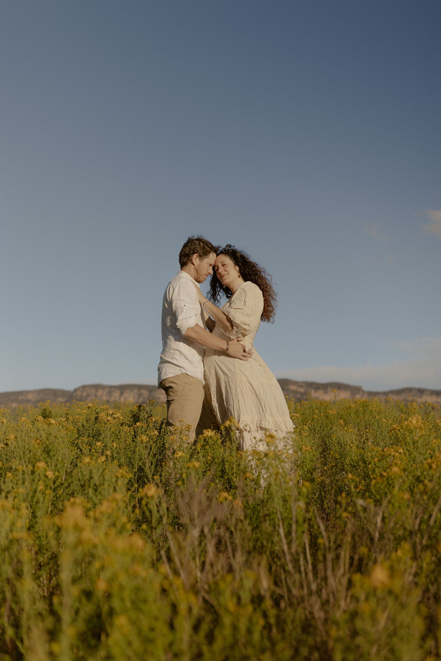 Intimate moment shared between couple standing in golden meadow with distant hills.