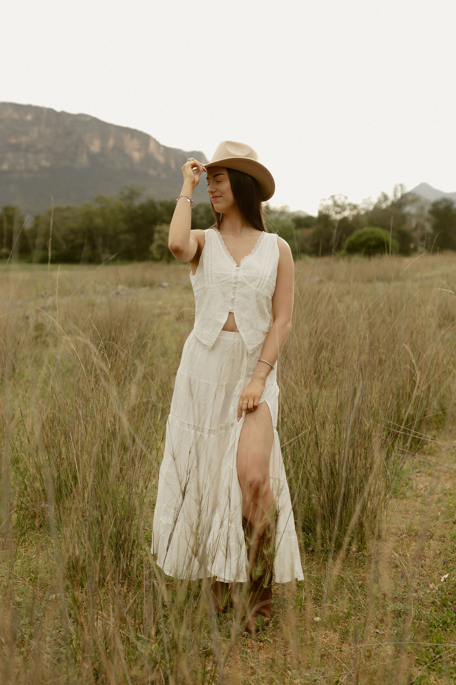 A person in a flowing white dress and sun hat walks through tall grass with mountains behind.