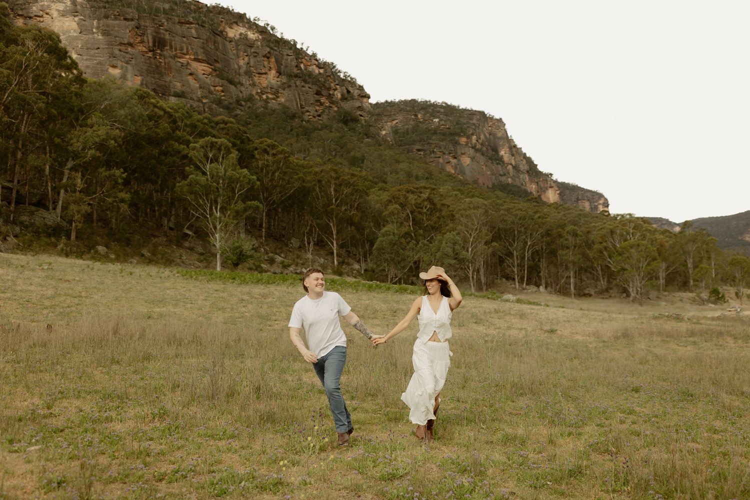 Two people run hand in hand through a grassy meadow with dramatic mountain cliffs rising in the background.