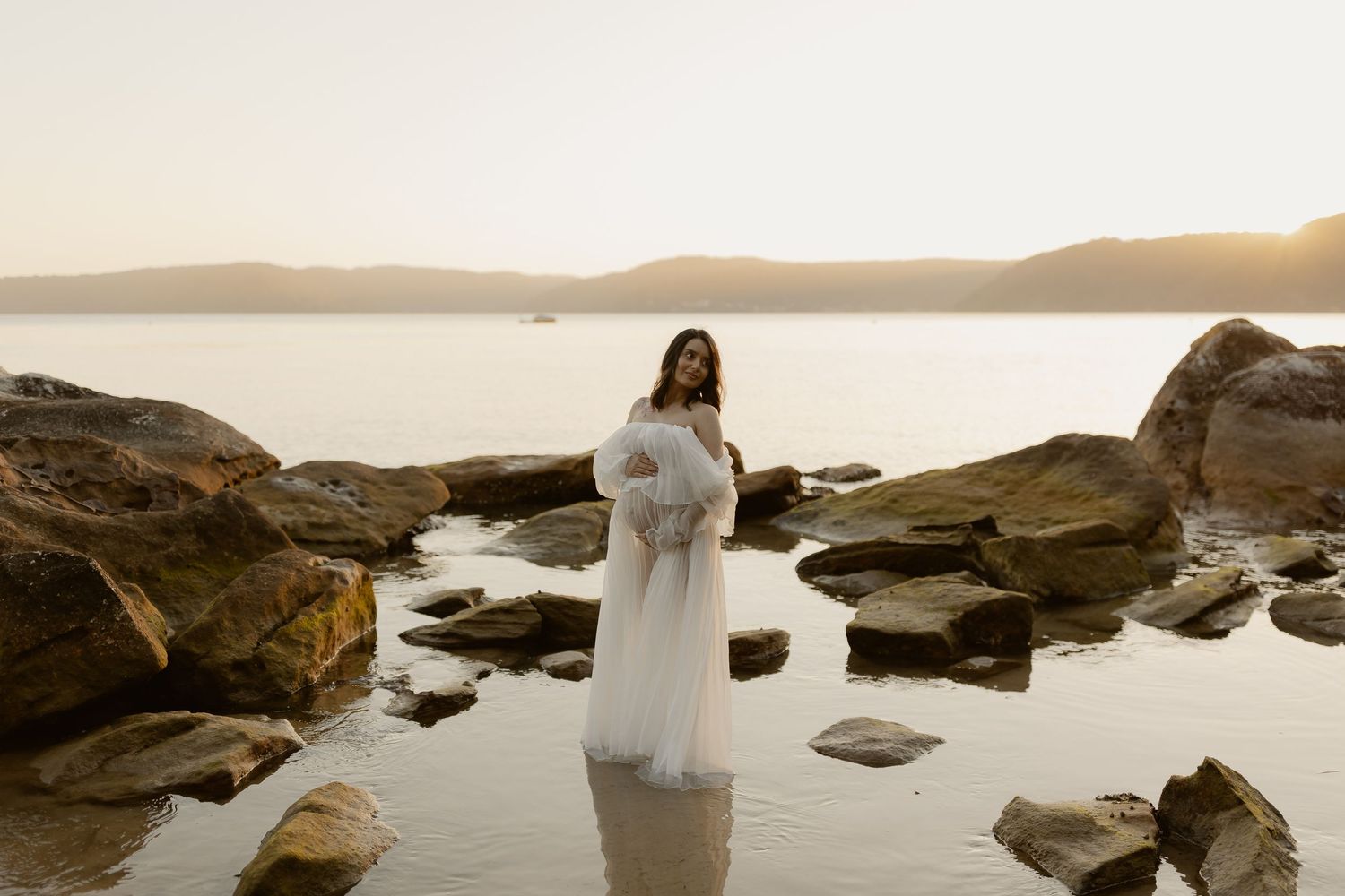 A romantic couple in wedding attire poses on rocky coastal cliffs at sunset with dramatic ocean views.