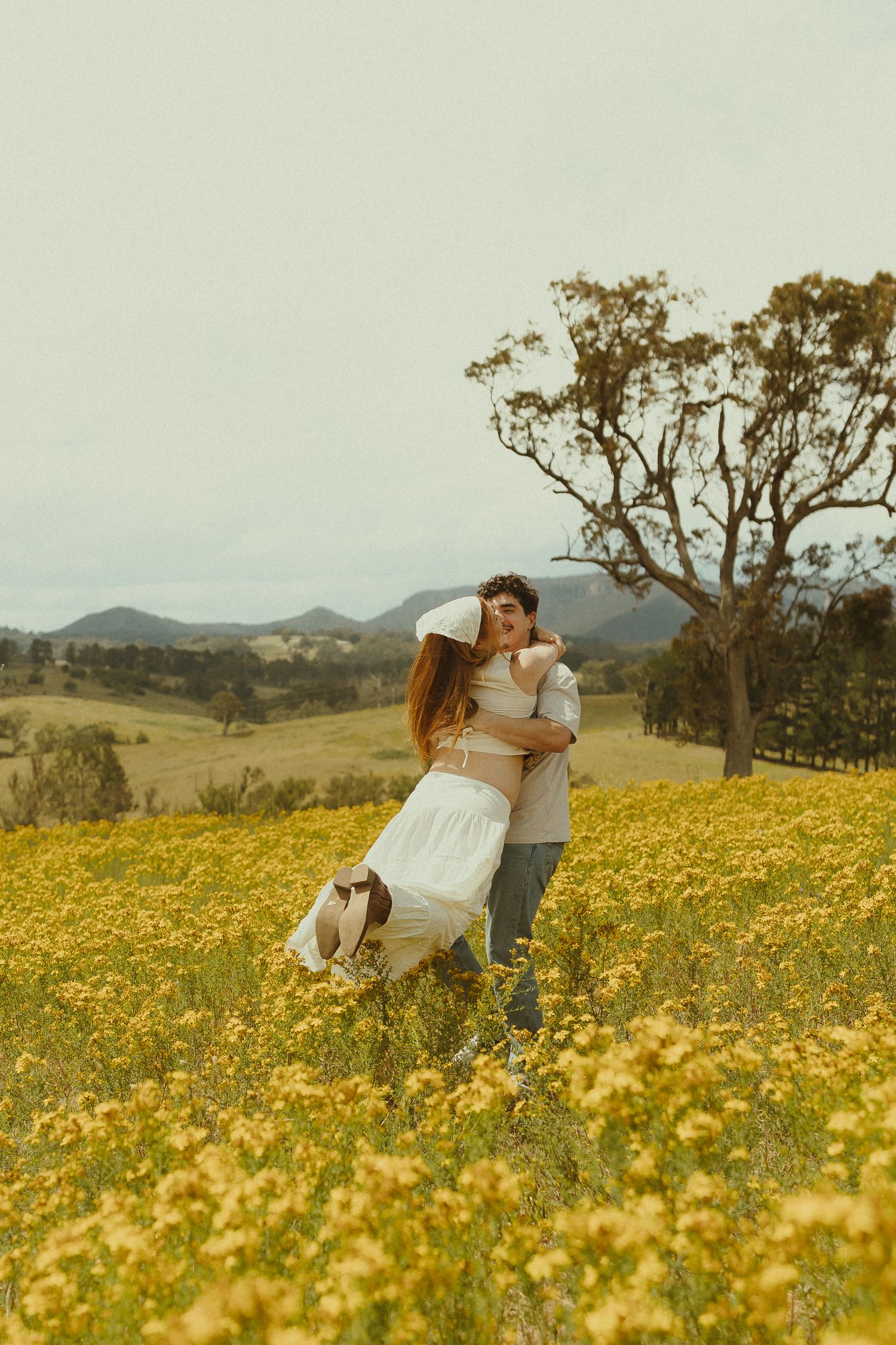 Couple embracing in a sunlit meadow of yellow wildflowers with rolling hills and trees in the background.