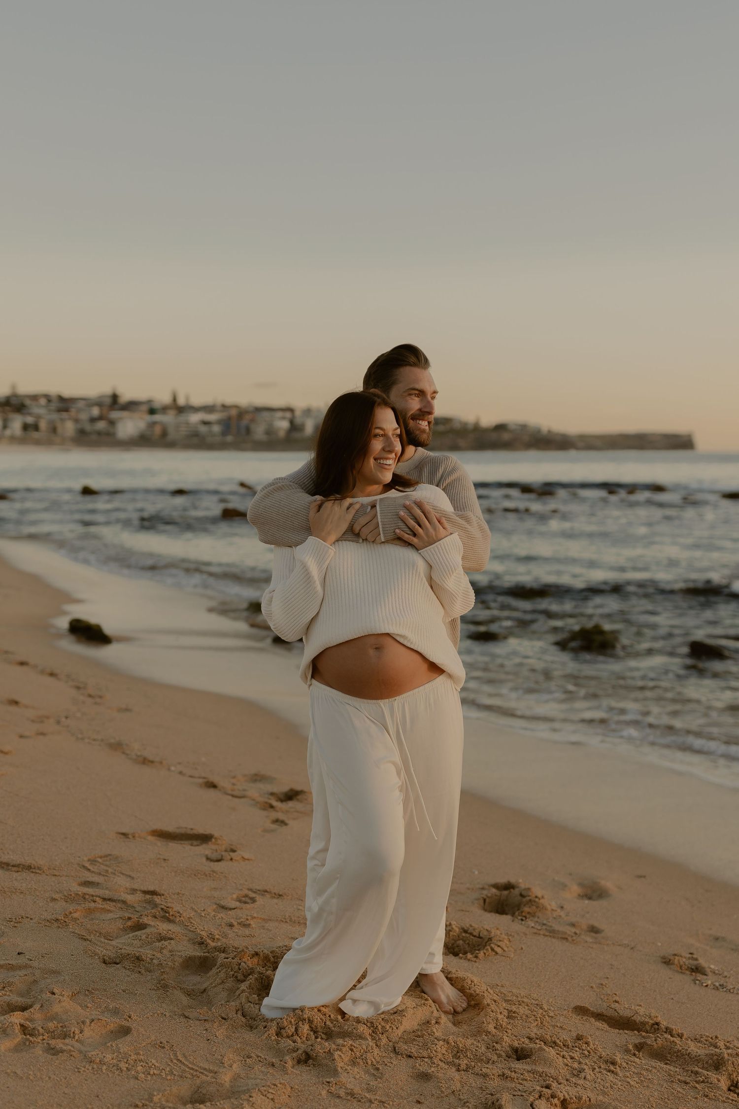 A romantic maternity photoshoot on the beach at sunset with a couple embracing in white outfits.