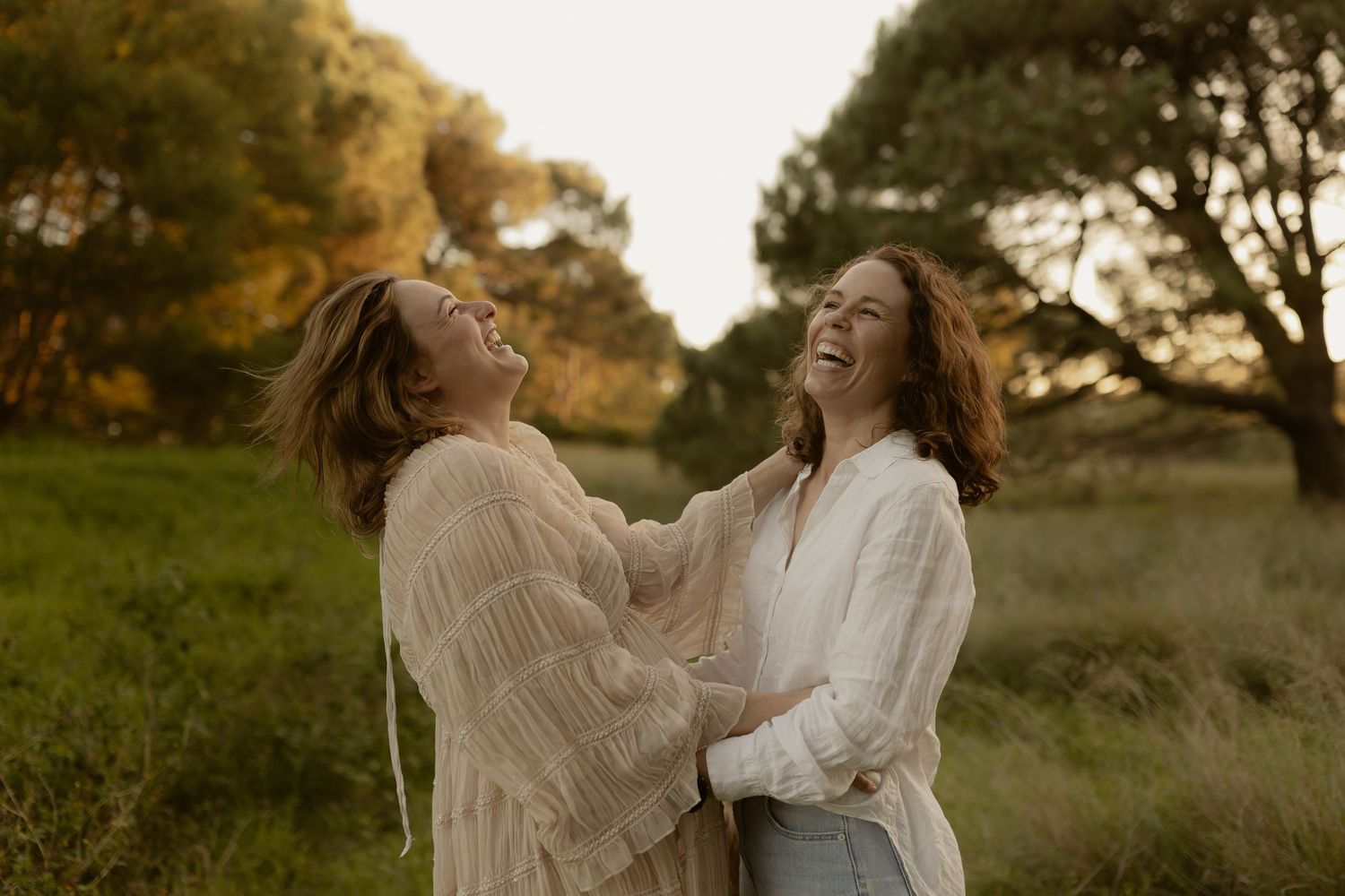 Two people in white sweaters laugh joyfully together in a golden autumn field during sunset.
