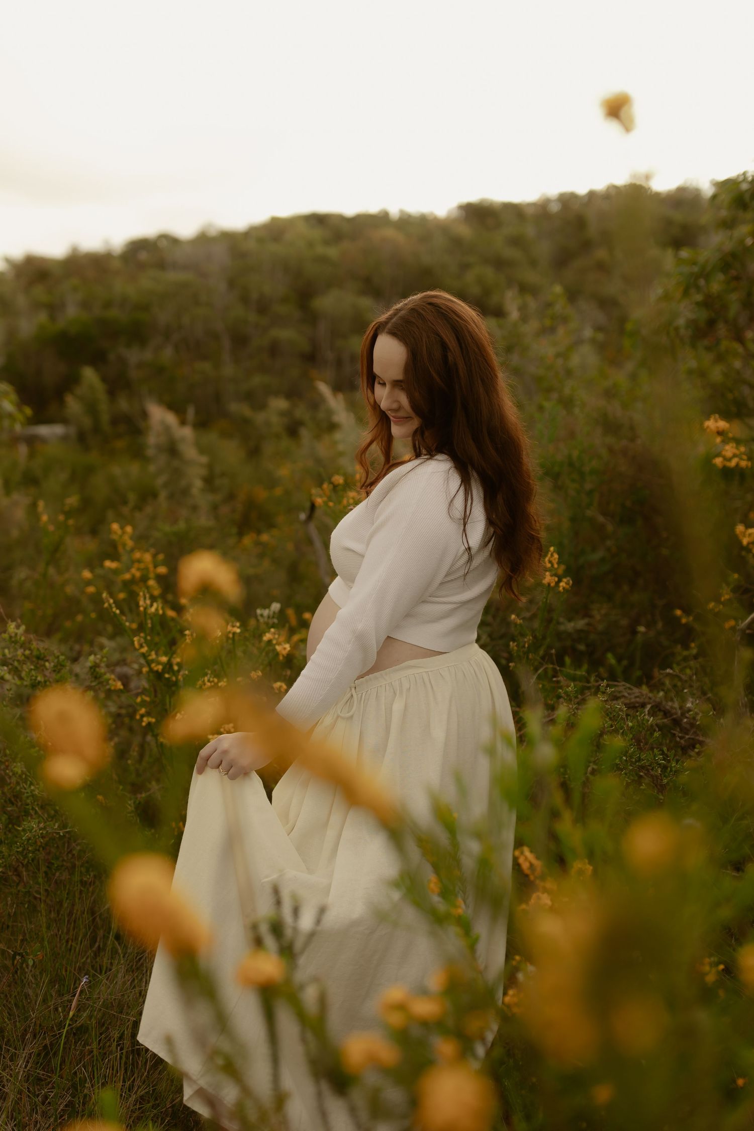 A woman in a white dress stands among yellow wildflowers in a lush green field at golden hour.
