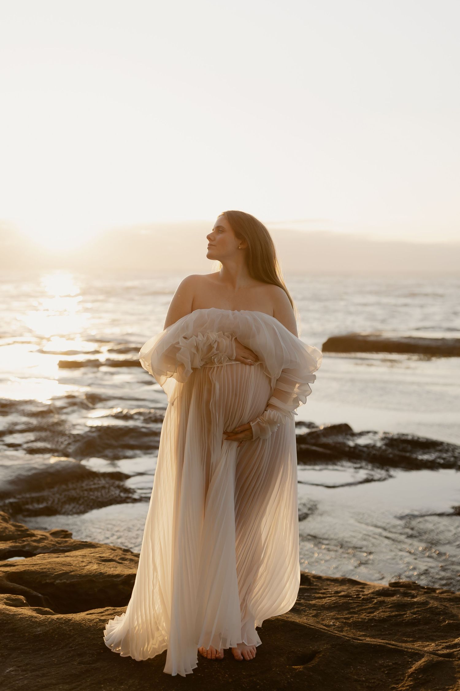 Ethereal off-shoulder cream gown with flowy pleats photographed on rocky coastline at golden hour sunset.