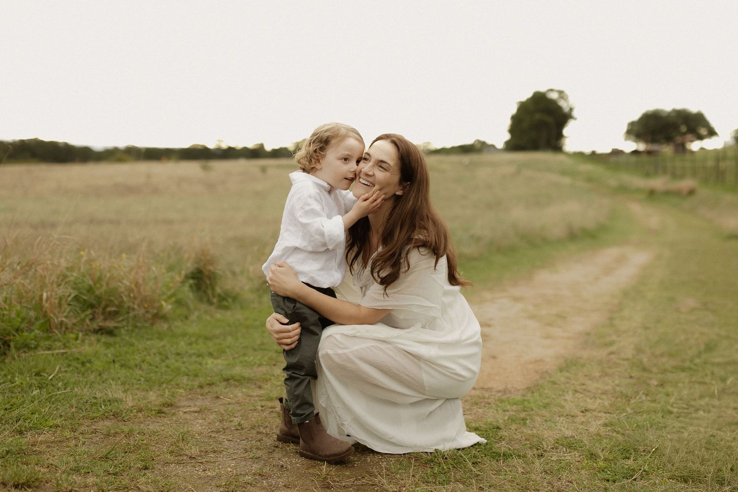 A couple in white outfits poses together in a grassy field at sunset.