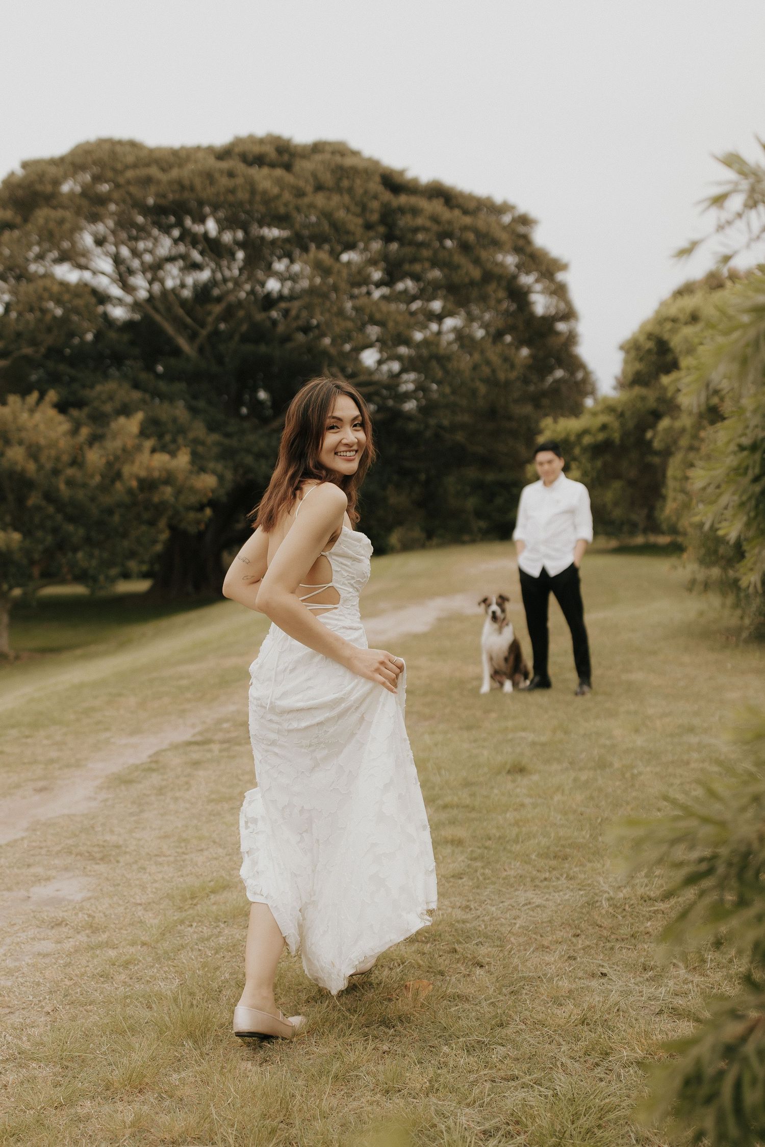 A bride in white dress runs playfully across a grassy hillside during a wedding photoshoot.