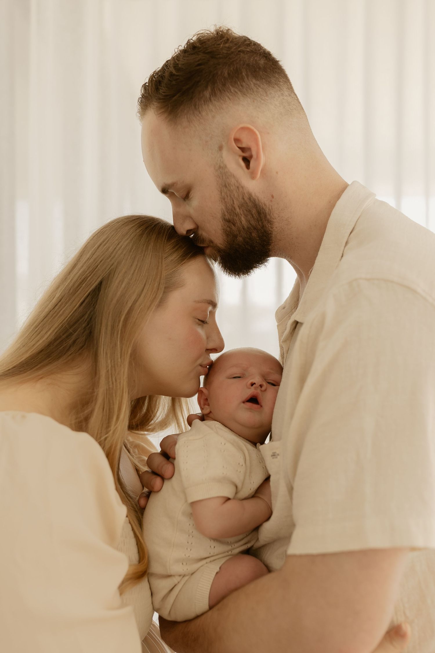 A touching family moment as parents in cream-colored outfits share an intimate moment with their sleeping newborn.