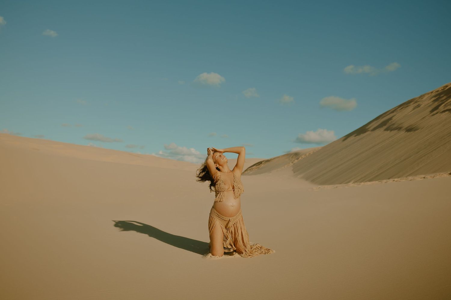 A person in flowing dress sits on golden sand dunes under a clear blue sky casting long shadows.