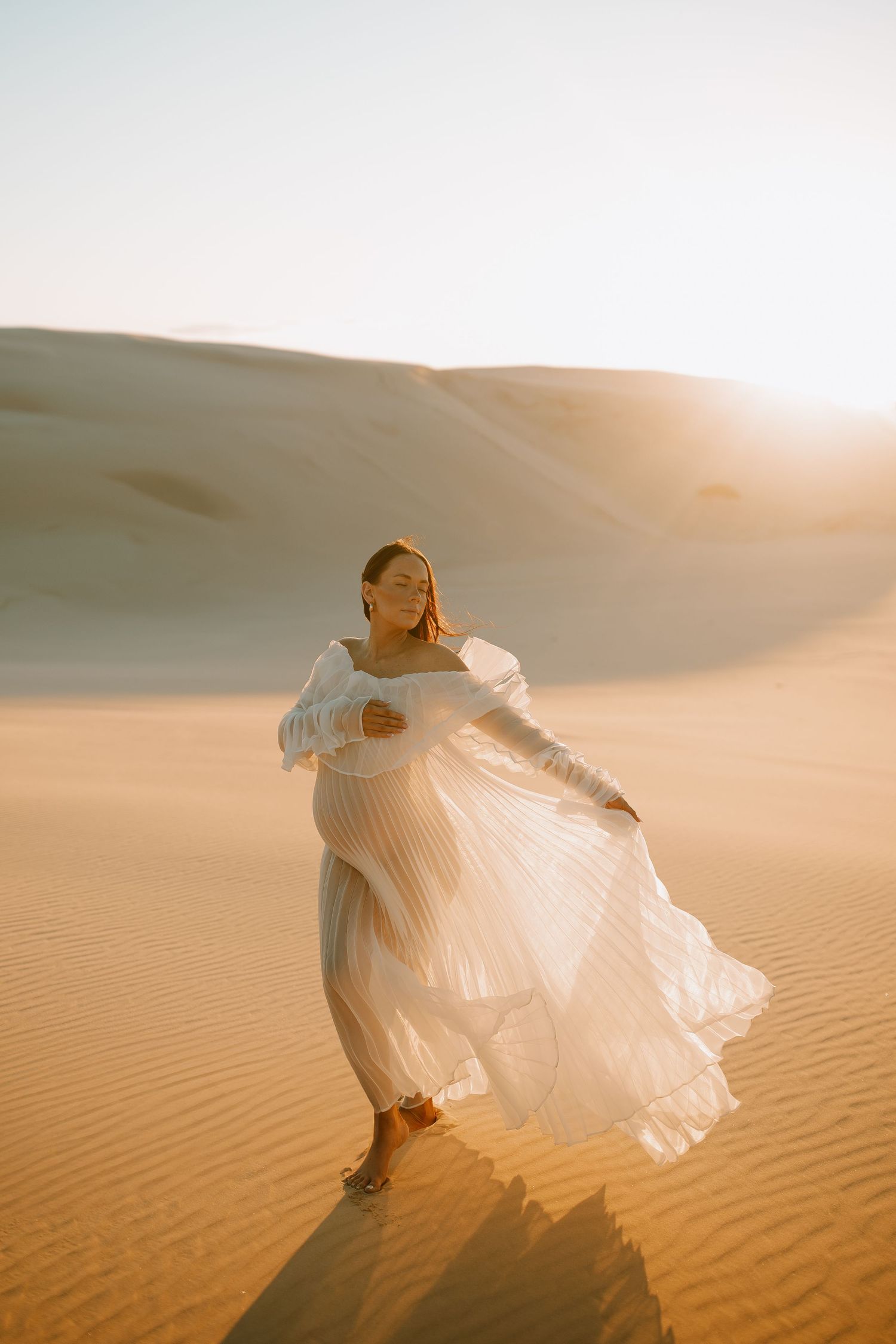 A person in a flowing white dress twirls gracefully on golden sand dunes during a sunset in the desert.