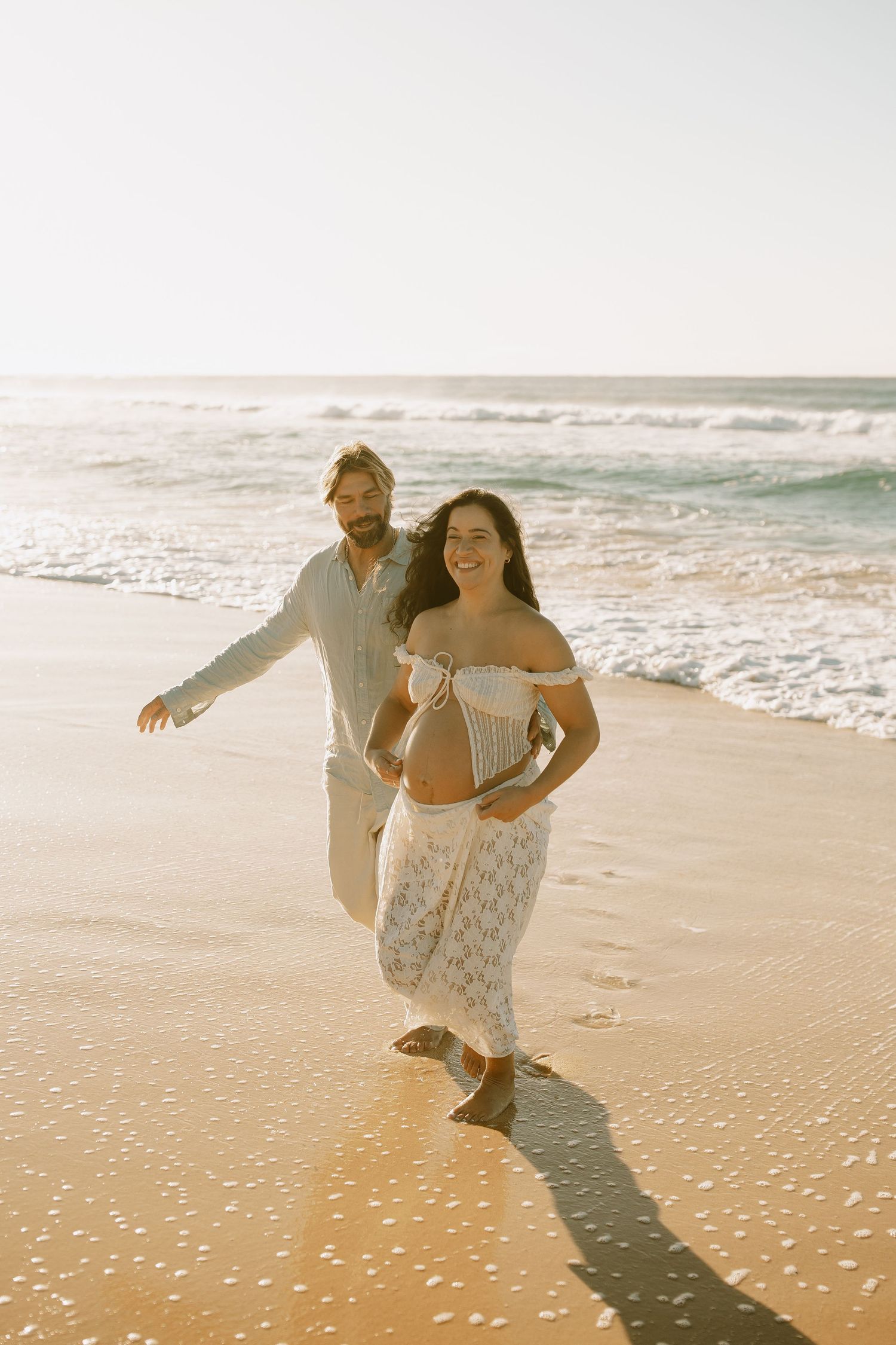 Carefree couple walking and laughing together on sunny beach with waves breaking on shore behind them.