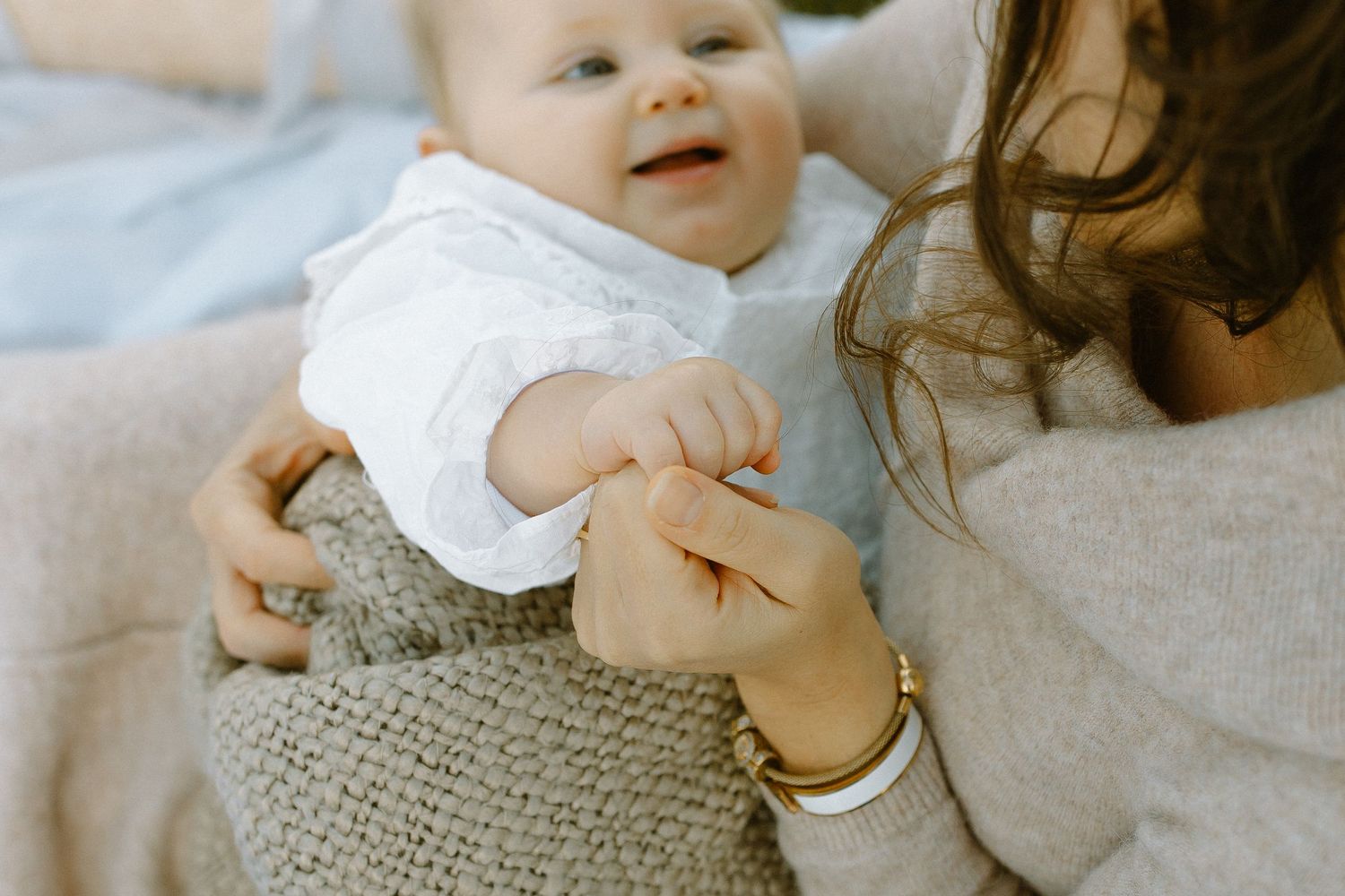 Infant in white outfit holds parent's hand during cozy indoor photoshoot.