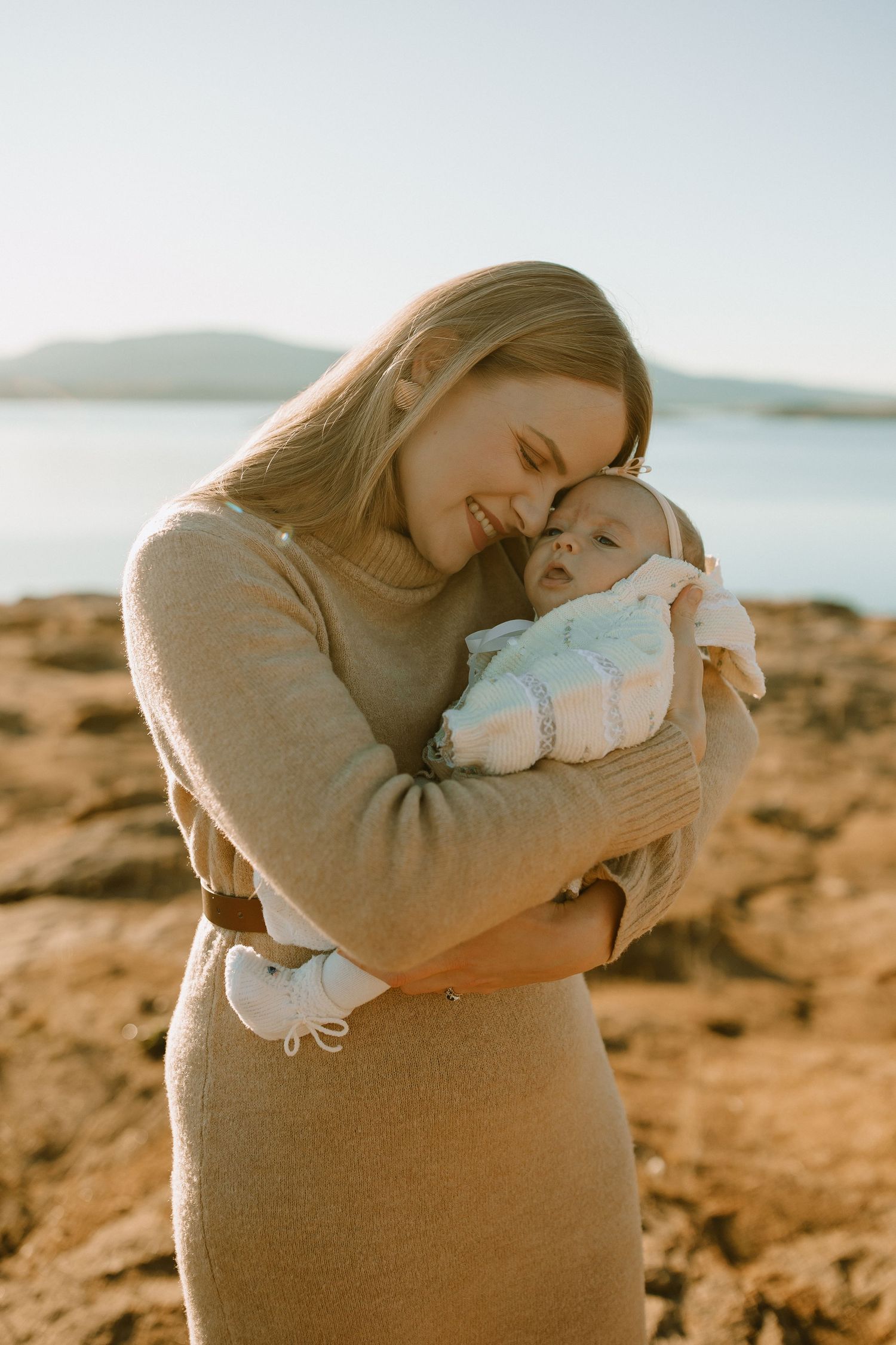 A person holding and smiling at their baby against a scenic coastal backdrop during golden hour.