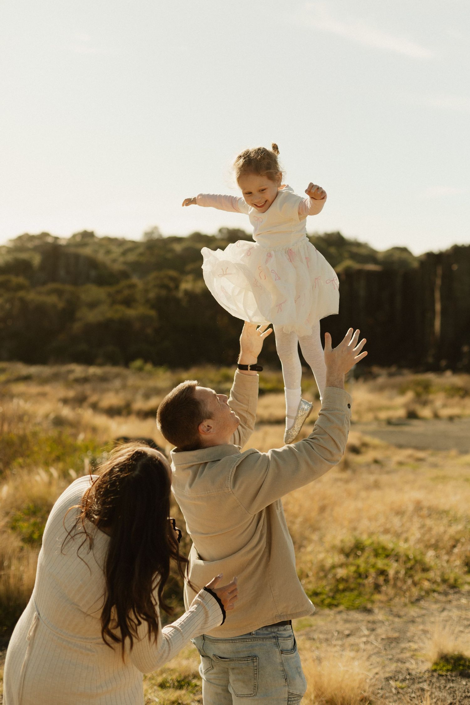 A joyful family moment as they lift and play with their child in a sun-drenched field.