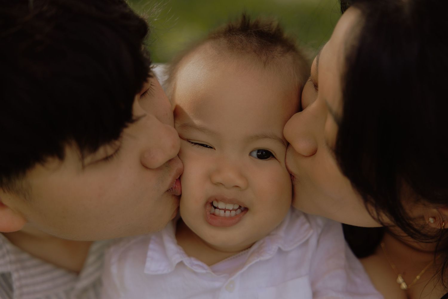 Two parents lovingly kissing their smiling baby on both cheeks in a tender family portrait outdoors.
