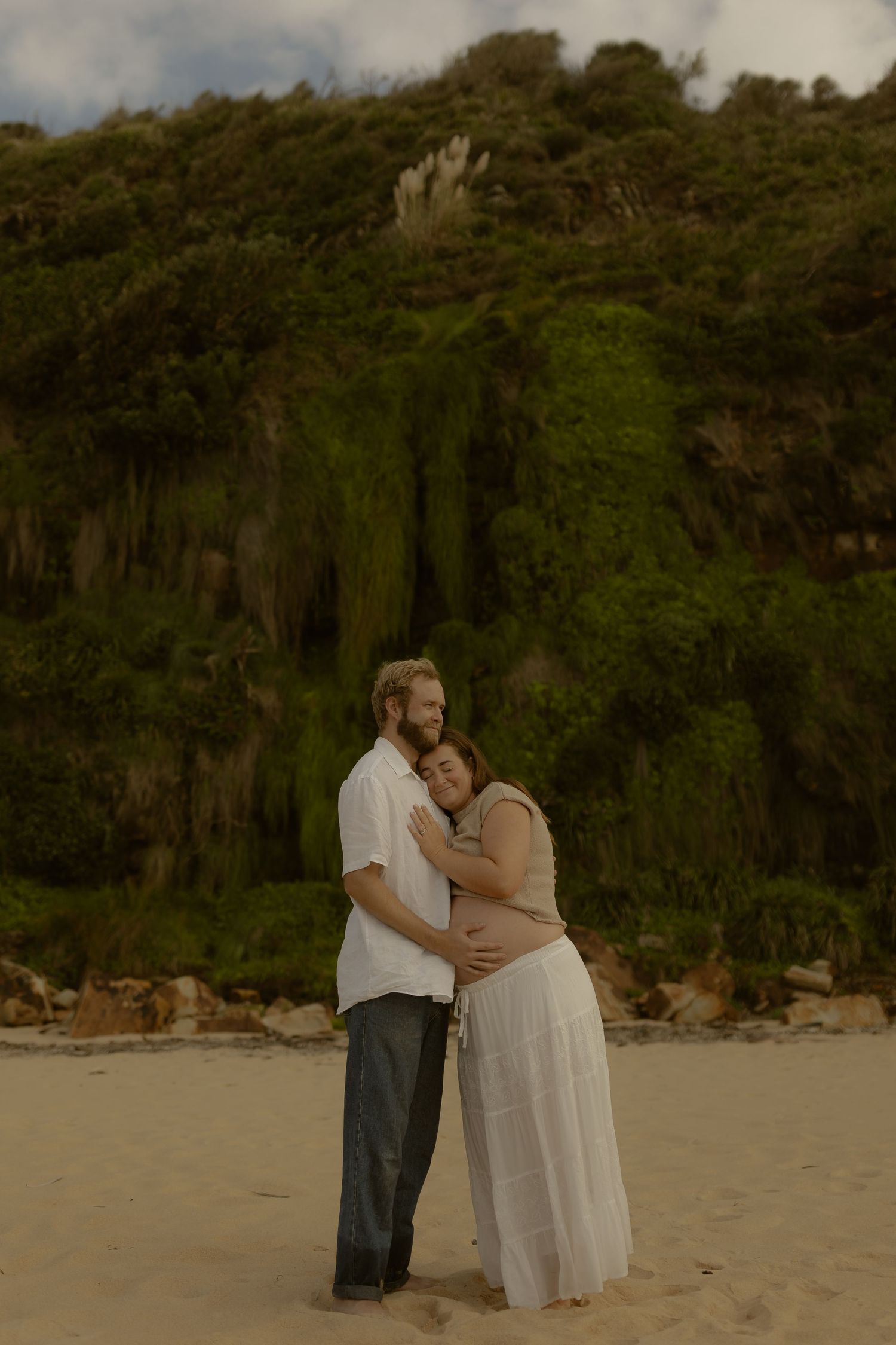 Two people embrace on a beach with lush greenery and cliffs in the background.