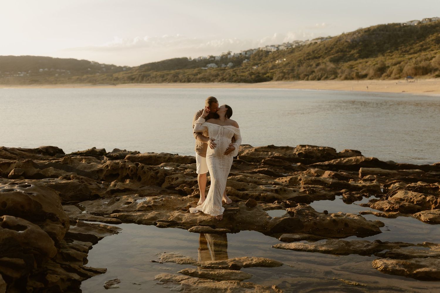 A couple stands together on rocky coastal cliffs overlooking the ocean at sunset creating a romantic silhouette.