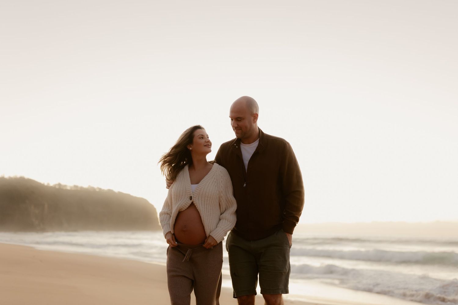 Couple walking on beach at sunset, pregnant woman cradling baby bump, man in black sweater, waves in background.