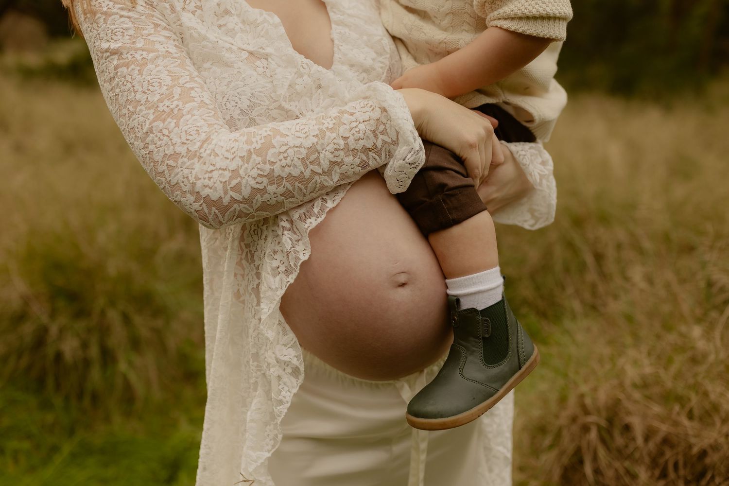 Close-up of pregnant belly with toddler being held, wearing green boots, outdoors.