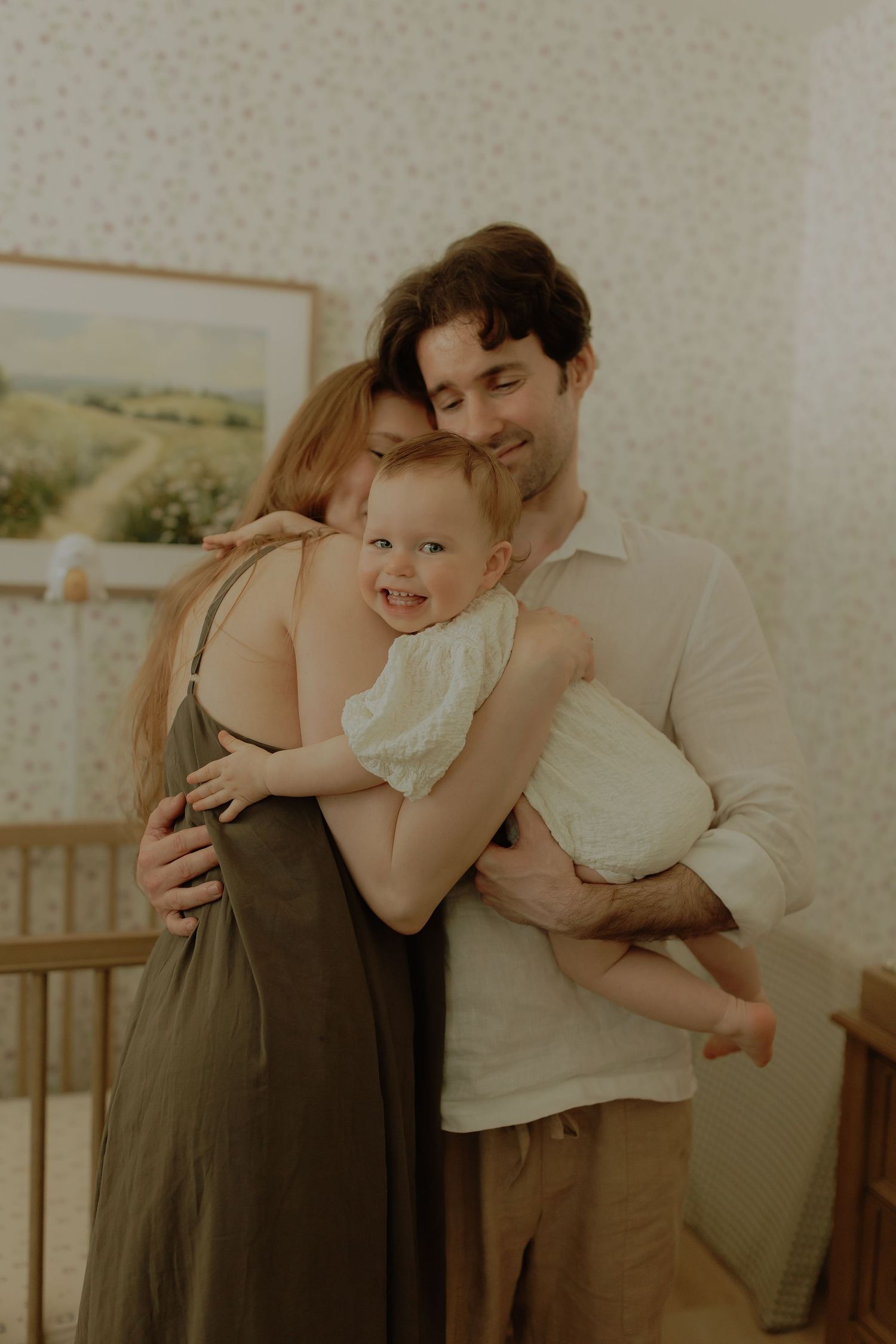 A smiling family of three shares a warm embrace in a cozy nursery bedroom.