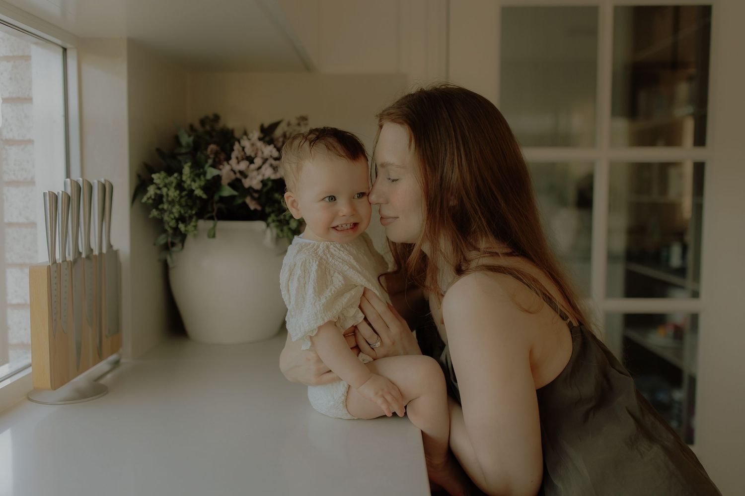A mother tenderly kisses her smiling baby while sitting in a bright, modern kitchen with white countertops.