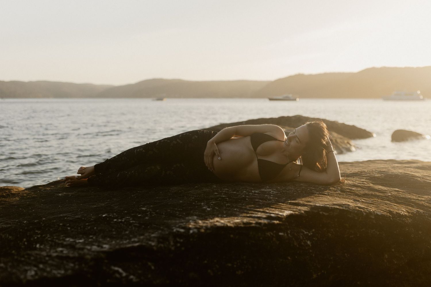 A figure rests on coastal rocks at sunset with calm ocean waters and distant hills in the background.