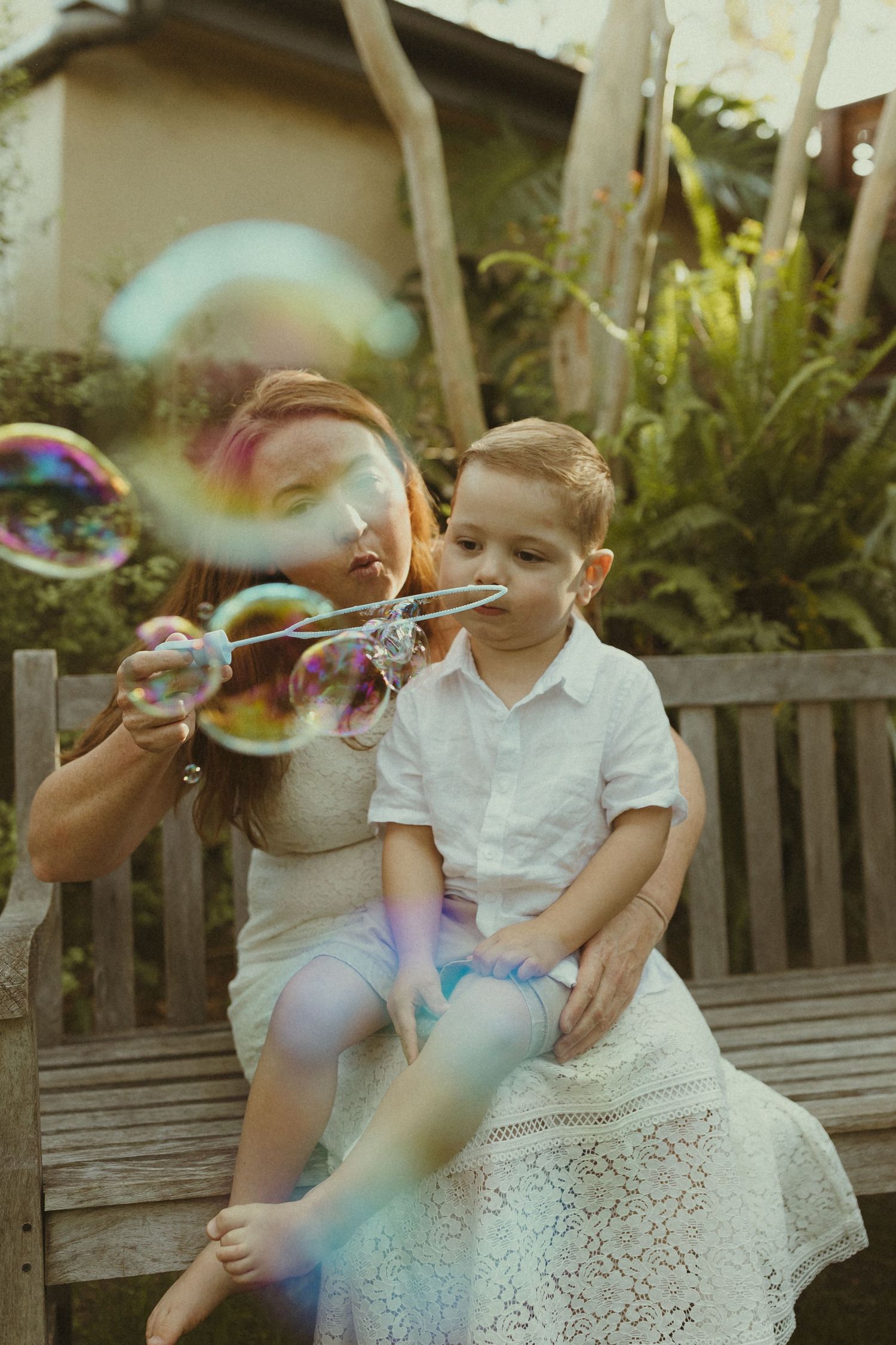 Children blowing bubbles while sitting on a wooden bench in a garden setting.