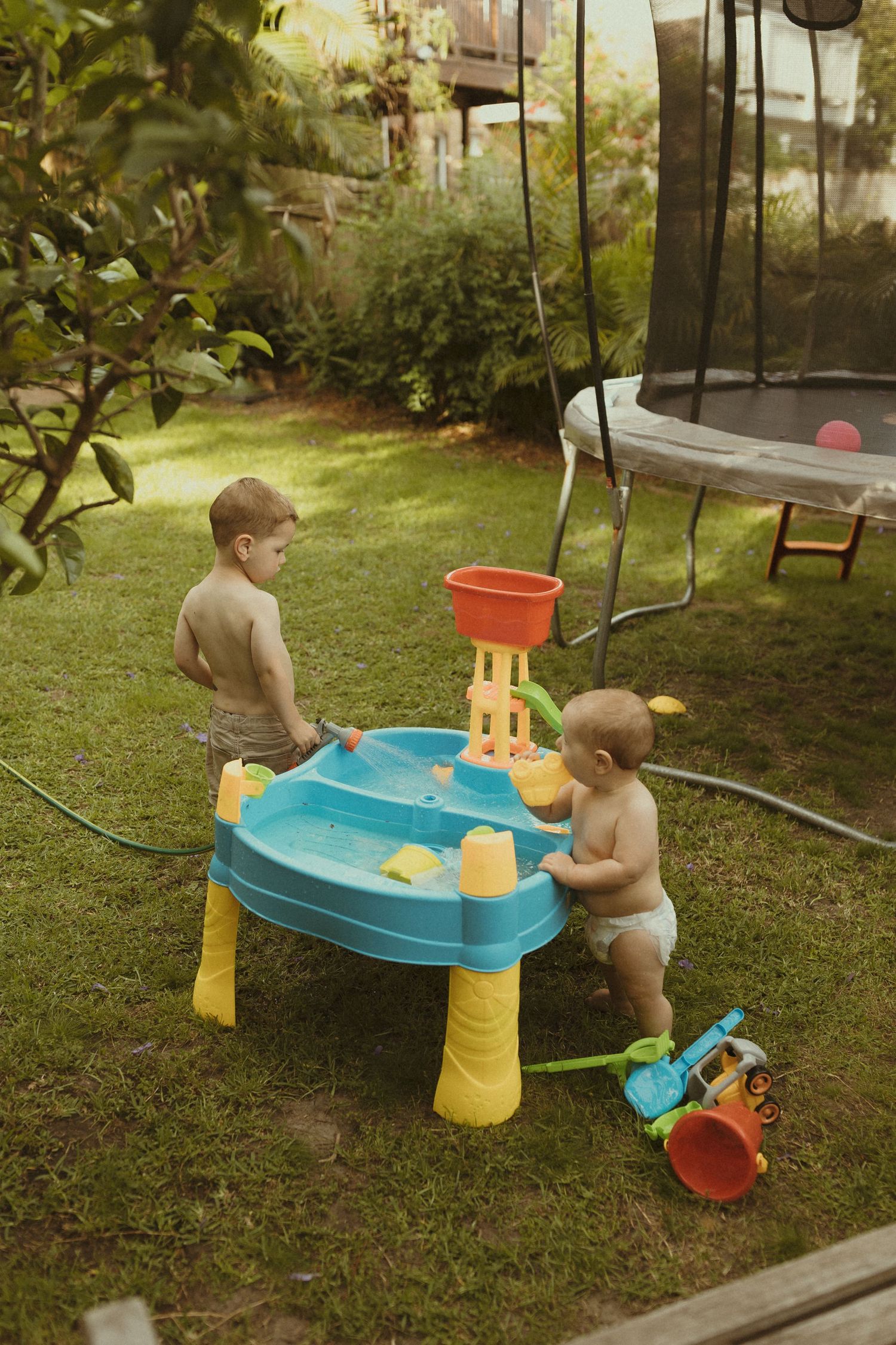 Children playing with a blue water table toy in a sunny backyard garden setting.
