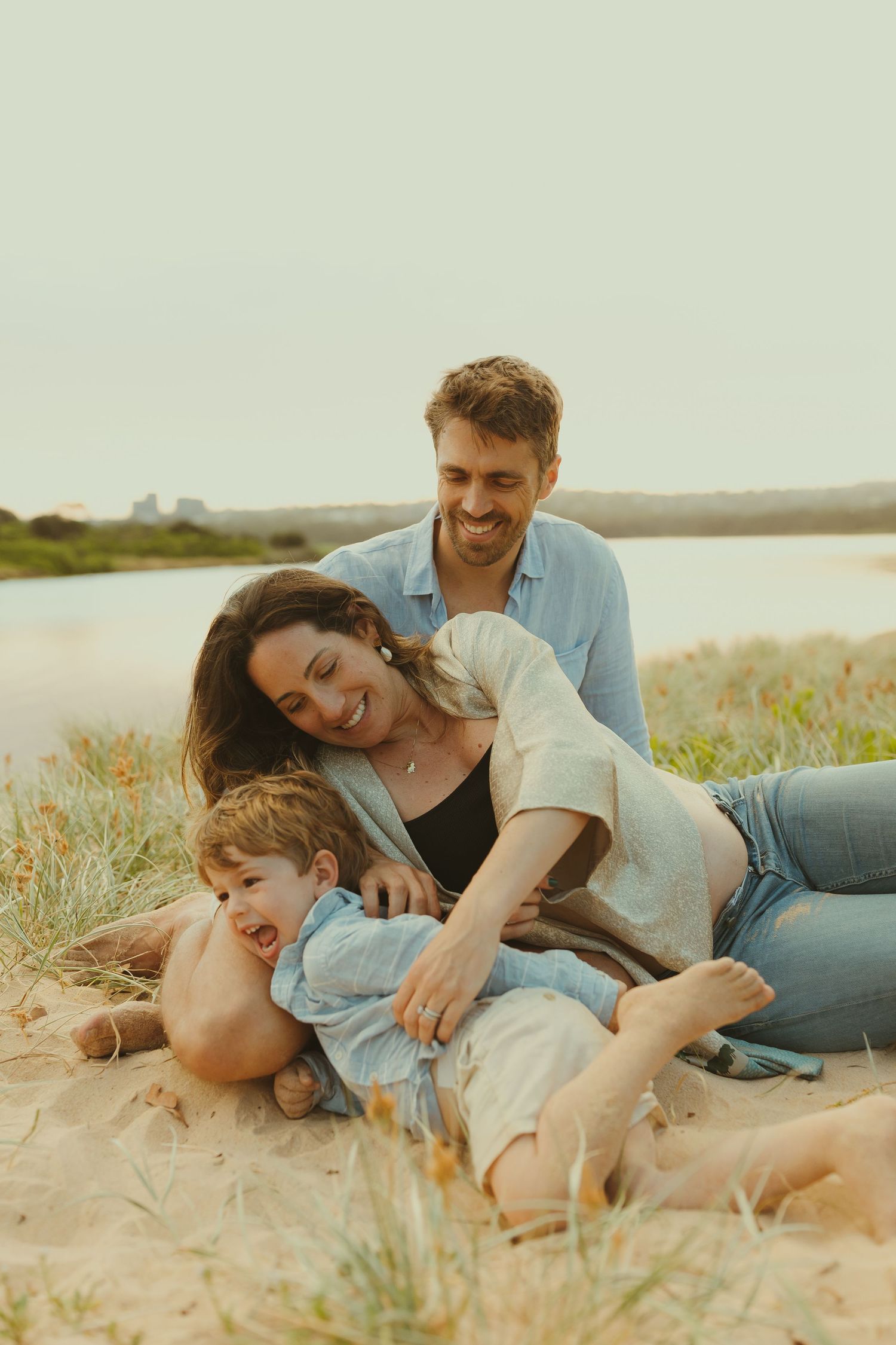 Family laying together playfully on sandy beach during golden hour photoshoot.