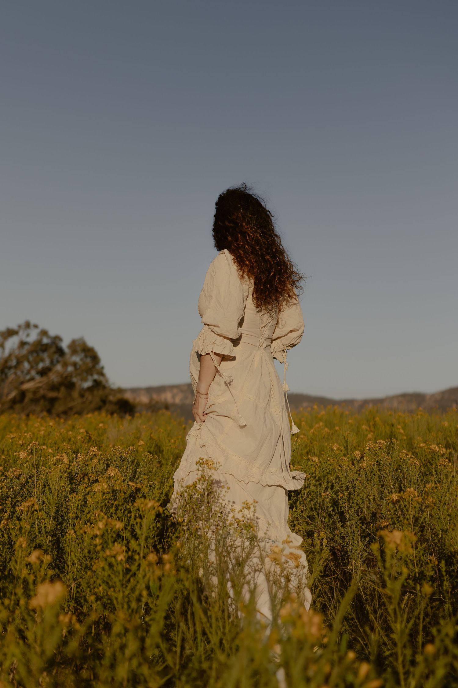 A person in a flowing cream dress stands alone in a field of yellow wildflowers at dusk.