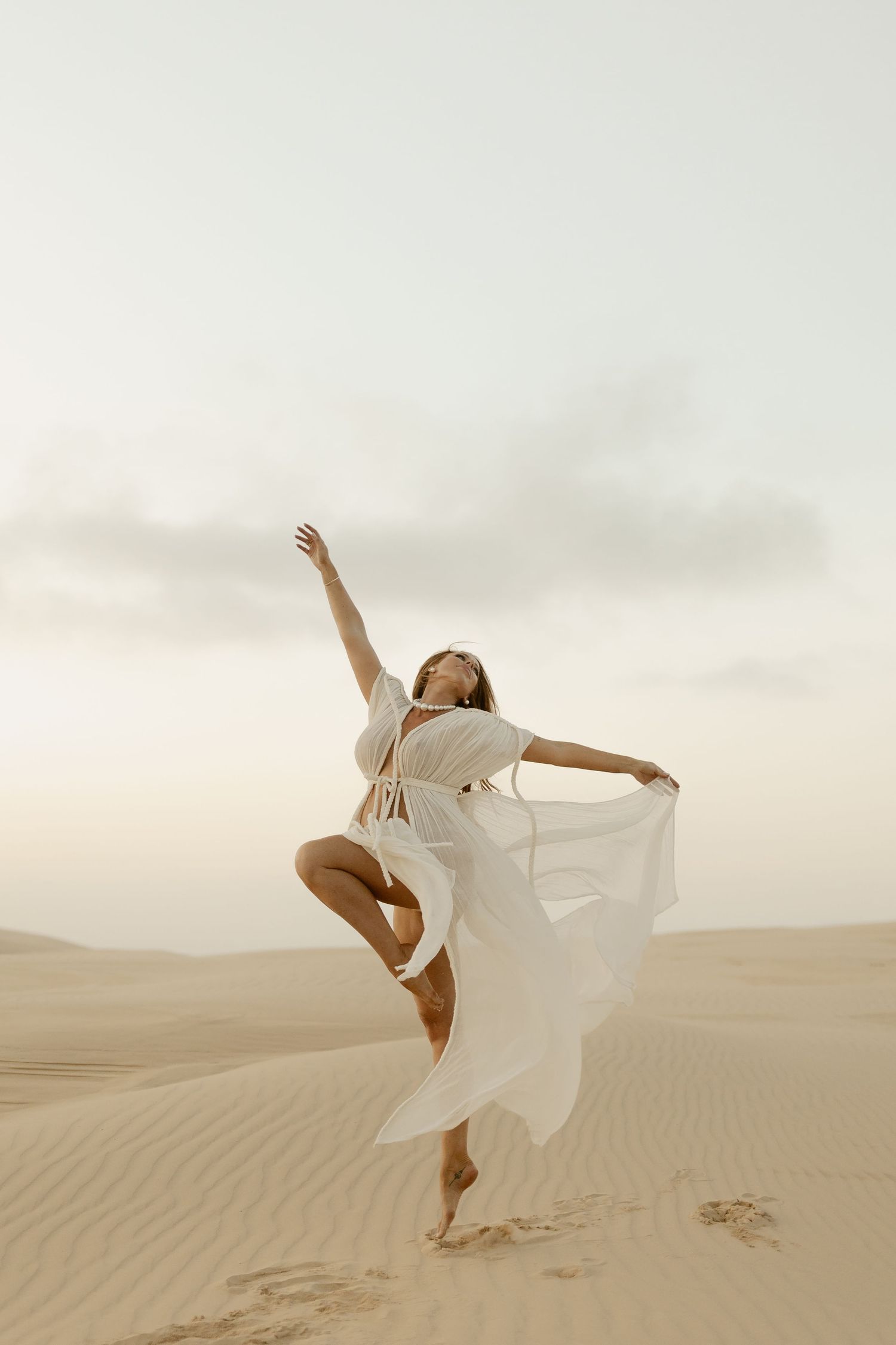 A figure in a flowing white dress dances gracefully across desert sand dunes at dusk.