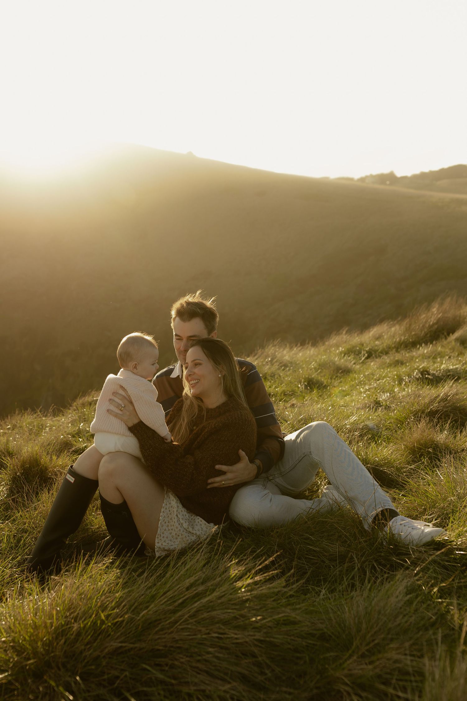 A couple sits together on a grassy hillside at golden hour, lovingly holding their baby.
