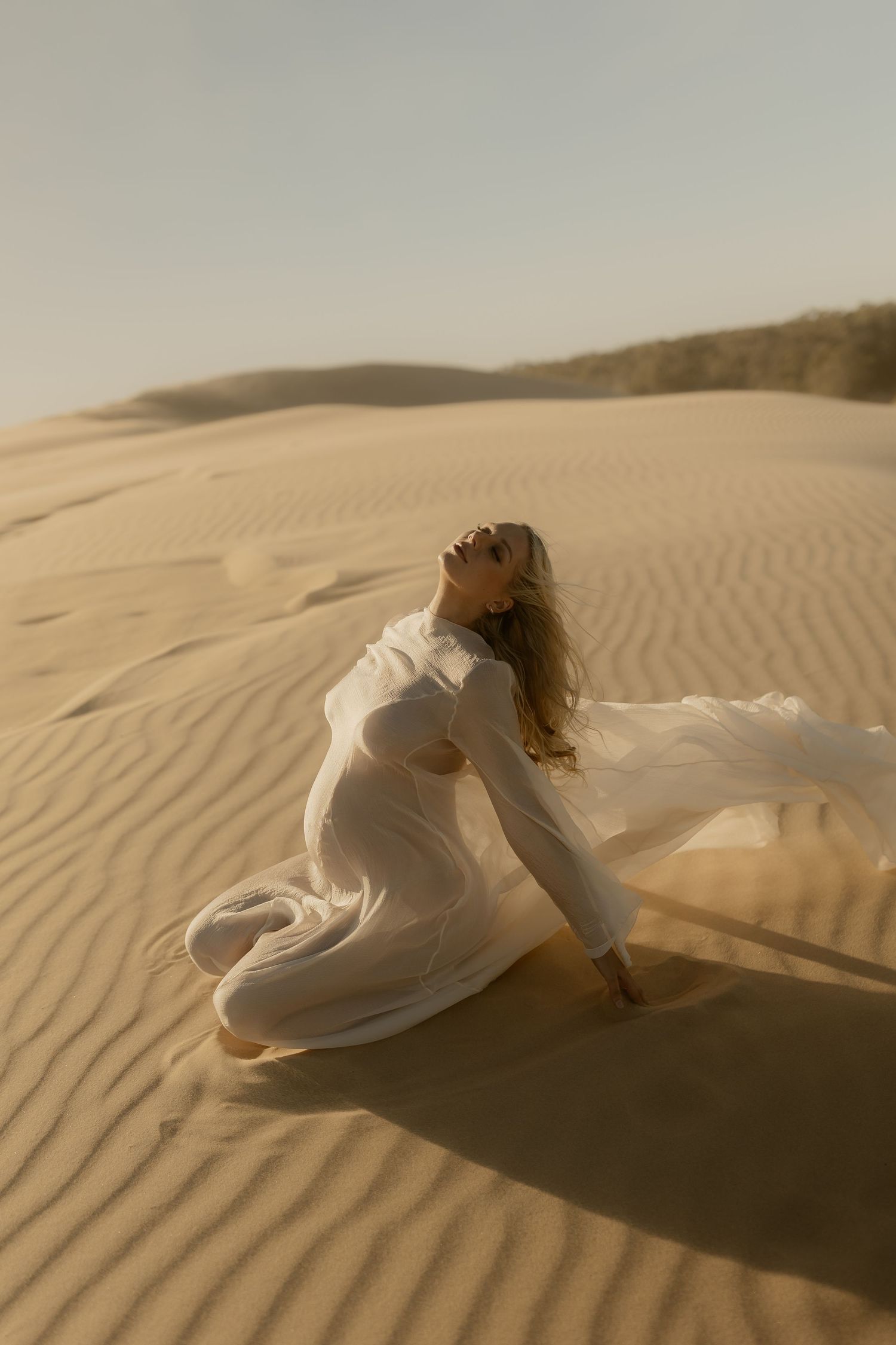 Woman in flowing white dress kneeling on golden sand dunes, hair blowing in wind, gazing upward in desert landscape.
