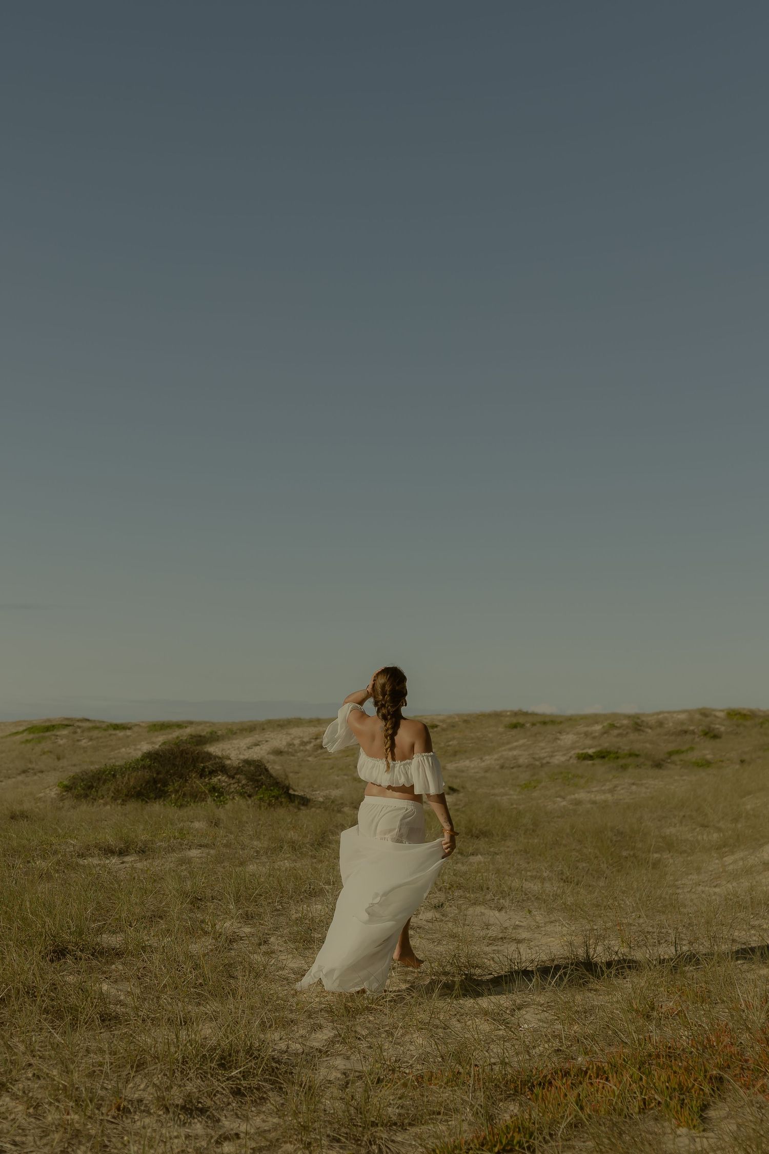 Woman in flowing white off-shoulder dress walks through sandy coastal dunes under a clear blue sky.