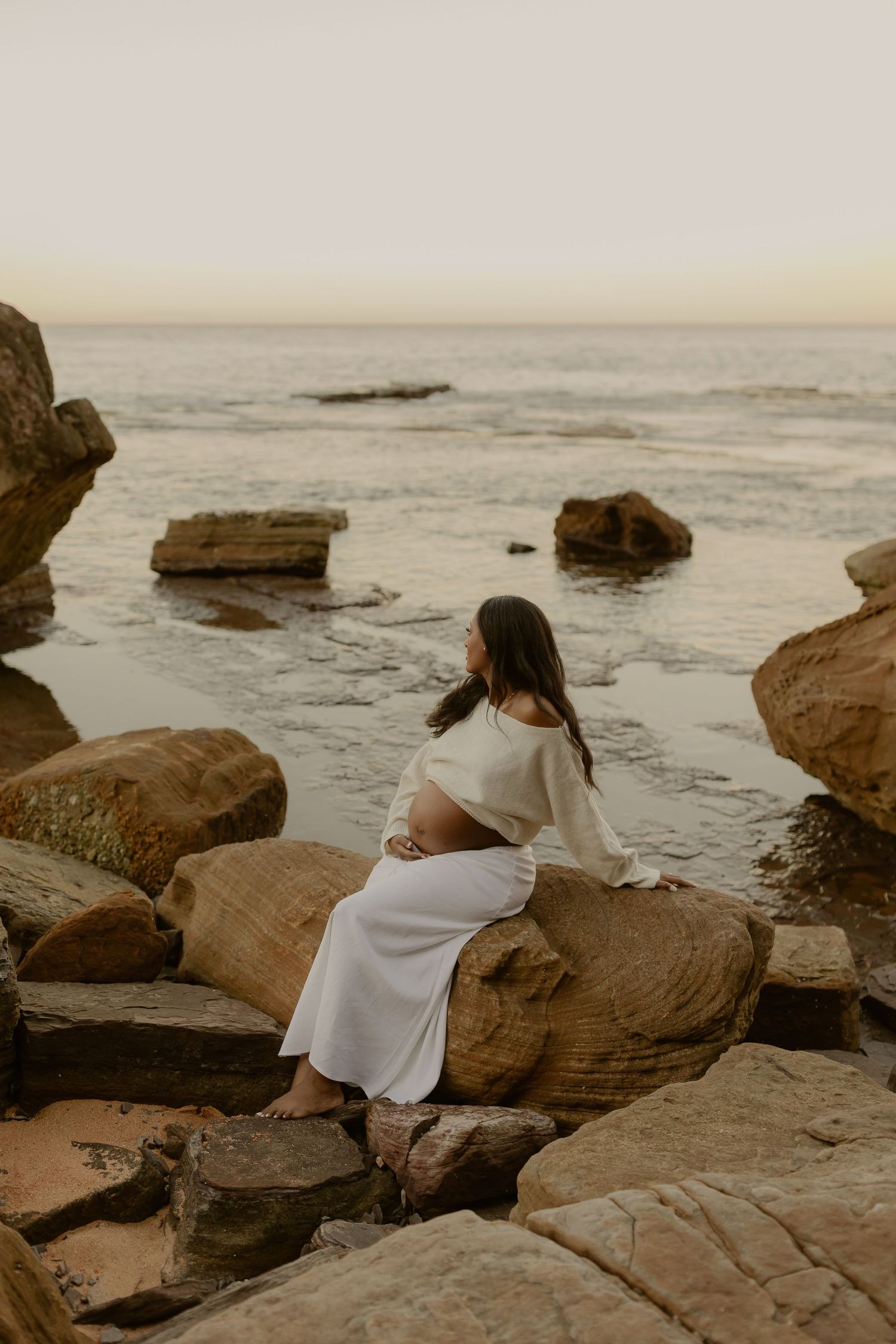 Pregnant woman in white outfit sits on coastal rocks at dusk, gazing at calm ocean during golden hour.