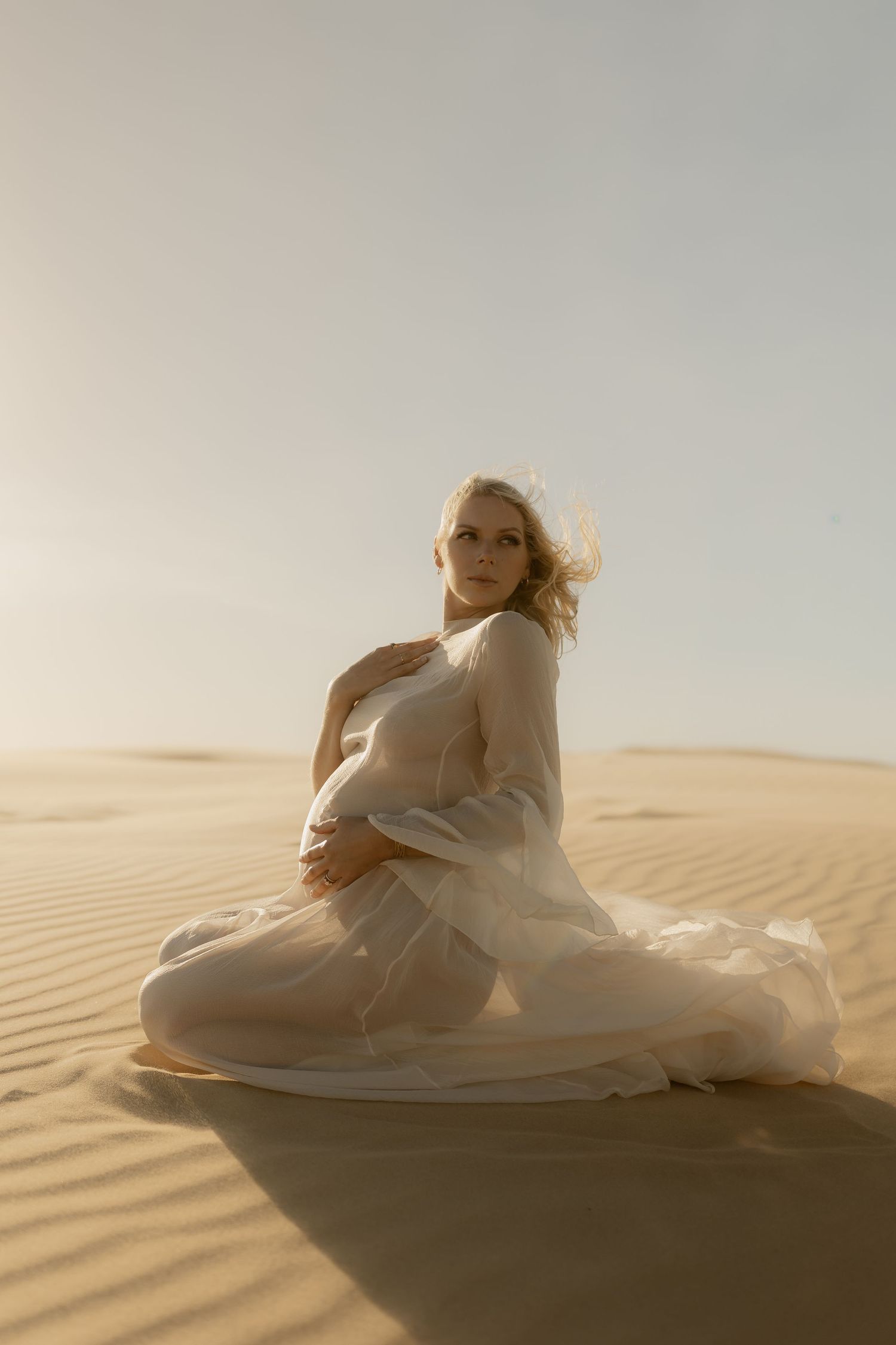 Pregnant woman in sheer white dress kneeling on golden sand dunes in soft sunlight.
